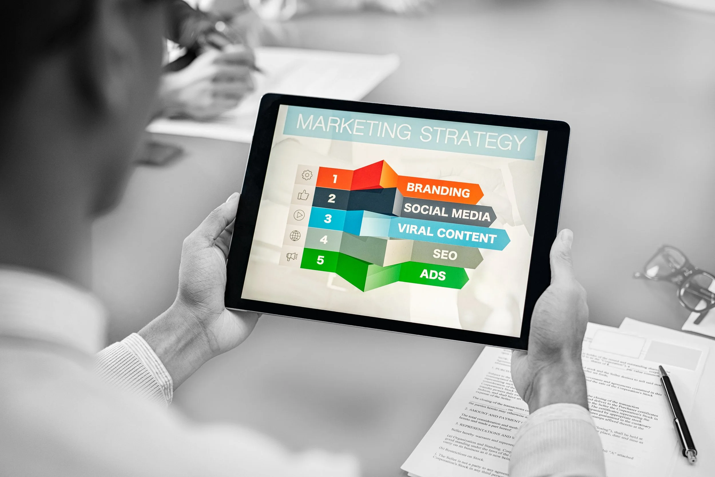 Person holding a tablet displaying a marketing strategy infographic with steps like branding, social media, viral content, SEO, and ads, in a professional meeting setting.