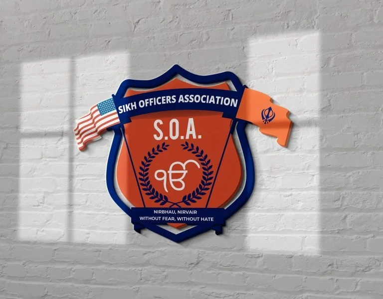 A badge or emblem for the Sikh Officers Association featuring the organization's name, the abbreviation S.O.A., an American flag and a Sikh flag, and the words 'Without fear, without hate' on a white brick wall background.