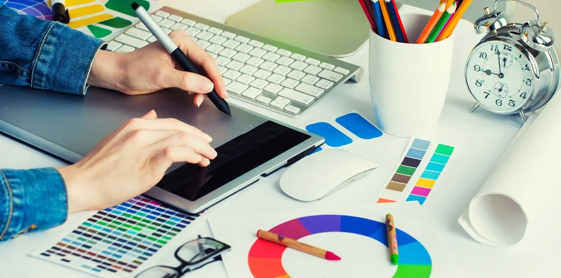 A designer working on a digital tablet at a white desk, surrounded by color swatches, a color wheel, a keyboard, graphic pens in a white cup, and an alarm clock.