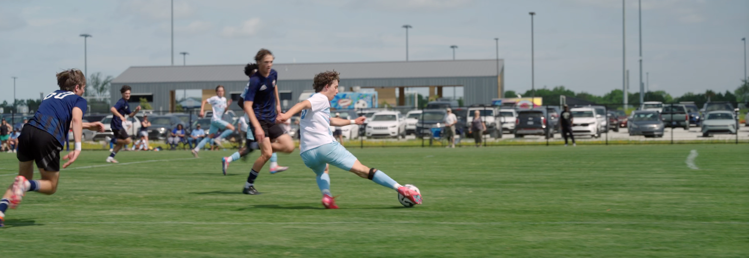 Young boys playing soccer on a field with a parking lot and building in the background.