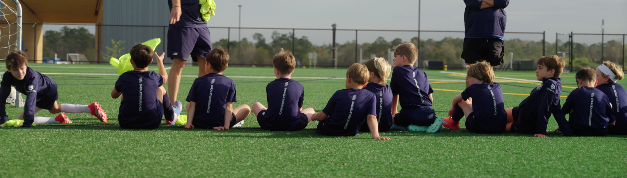A group of young children sitting on a soccer field, listening to coaches during a soccer practice.
