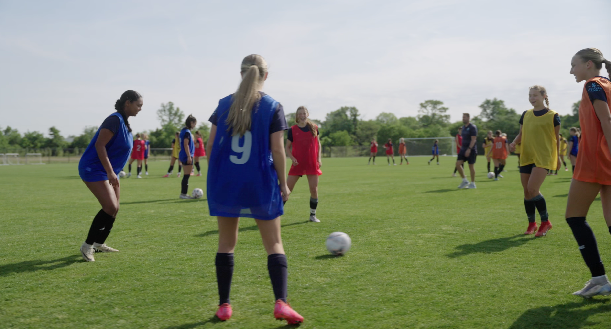 Group of young girls playing soccer on a grassy field, some wearing colorful jerseys and shorts, with trees and a clear sky in the background.