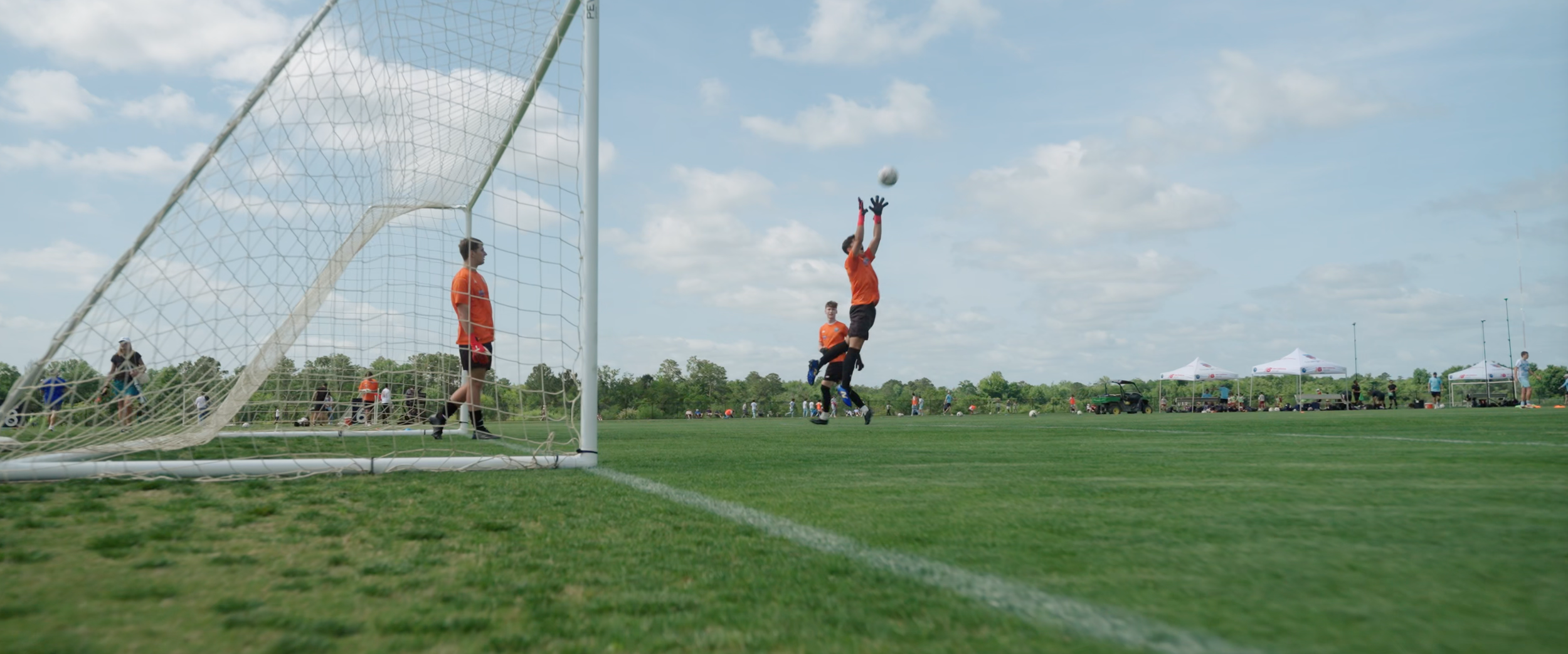 Soccer game with a goalkeeper in orange jumping to catch a ball near the goal, with another player in orange standing nearby and people in the background on a grassy field under a partly cloudy sky.