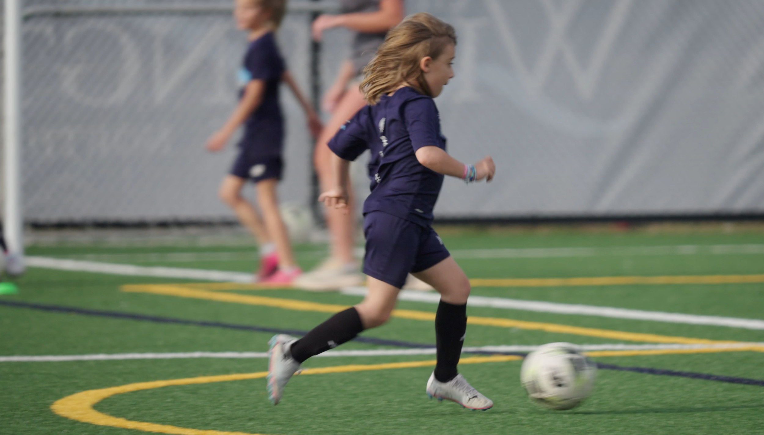 Young girl in sports uniform running with a soccer ball on an indoor turf field.