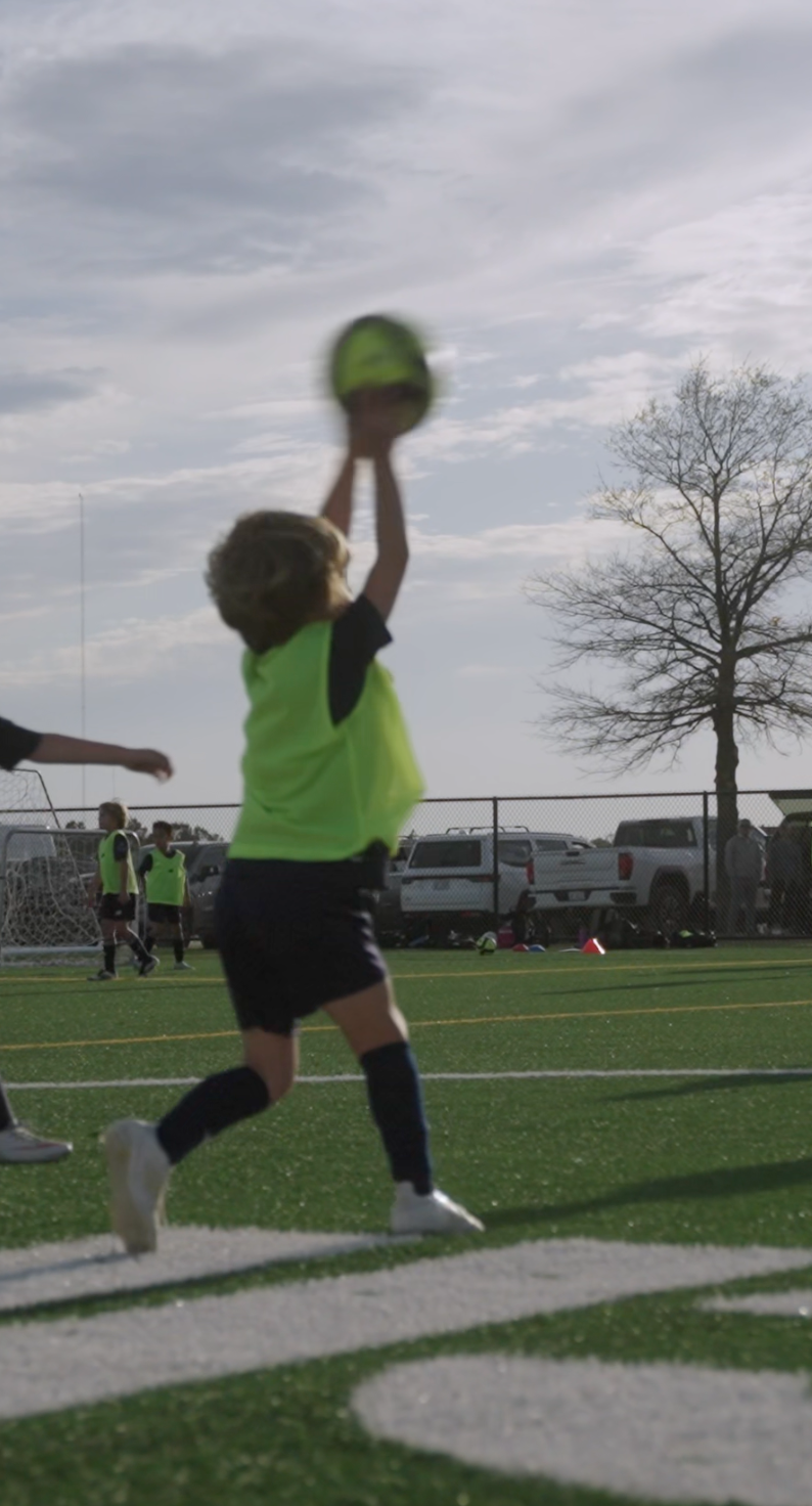 Child in neon yellow sports jersey reaching up to catch a football during a soccer game on a field.