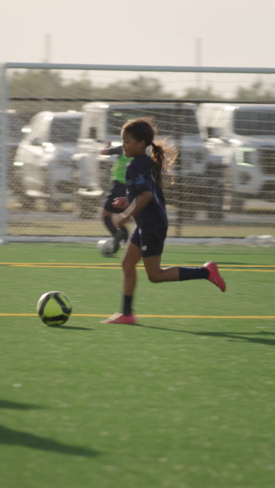 A young girl playing soccer on a field with a striped black and yellow ball, wearing pink shoes, dark shorts, and a dark sports jersey, with a fence and trucks in the background.