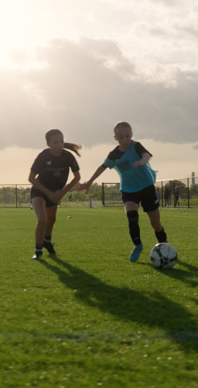 Two kids playing soccer on a field during late afternoon or early evening under cloudy sky.