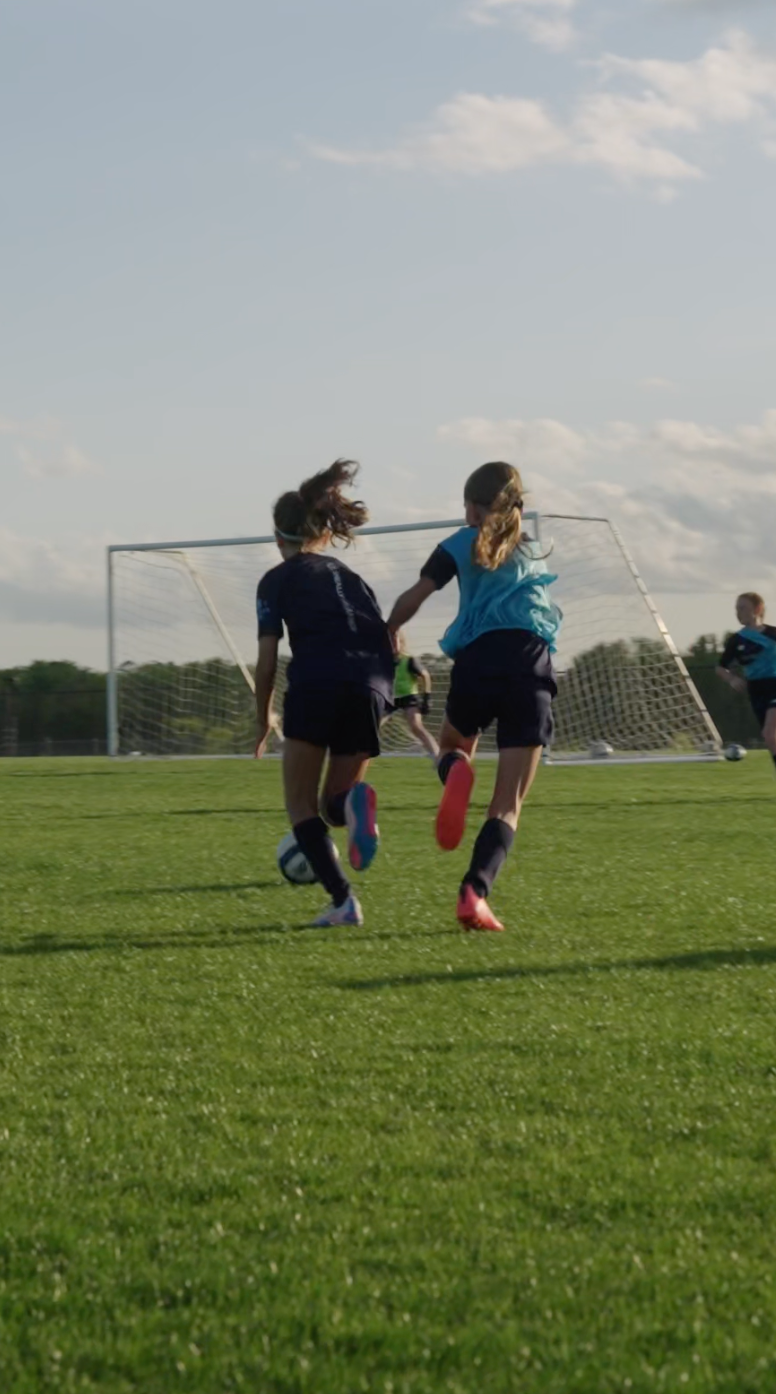 Two girls playing soccer on a green field with a goalpost in the background, under a partly cloudy sky.