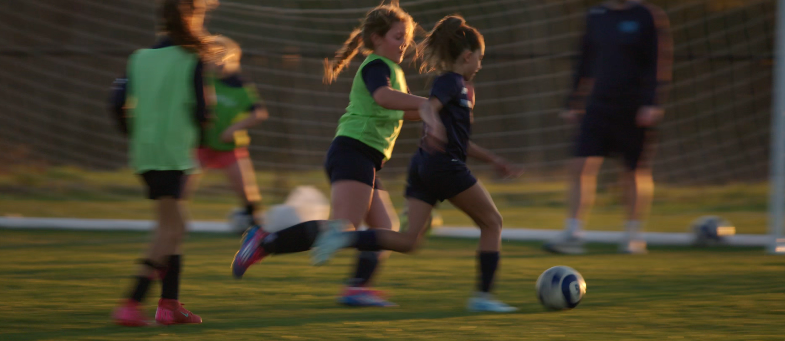Kids playing soccer on a field during sunset, with a focus on two girls running after the ball, wearing sports jerseys and shorts.