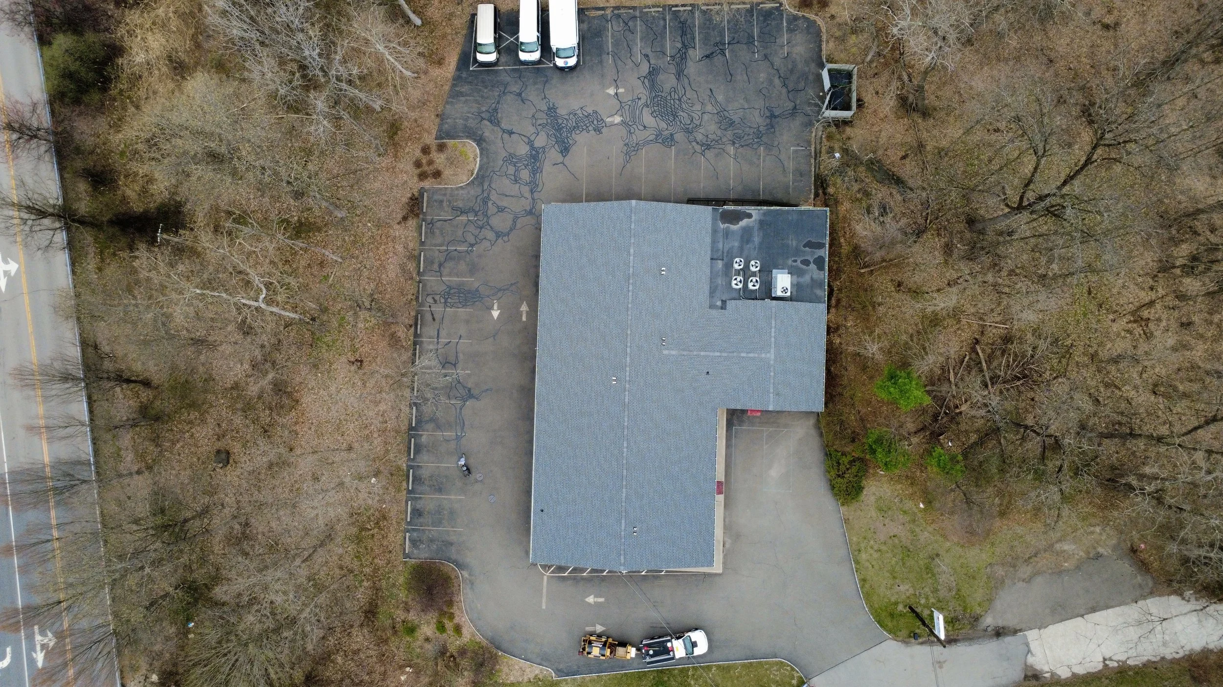 Aerial view of a parking lot with several cars, a building with a gray roof near trees, and a road with a turn, surrounded by trees and some grass areas.