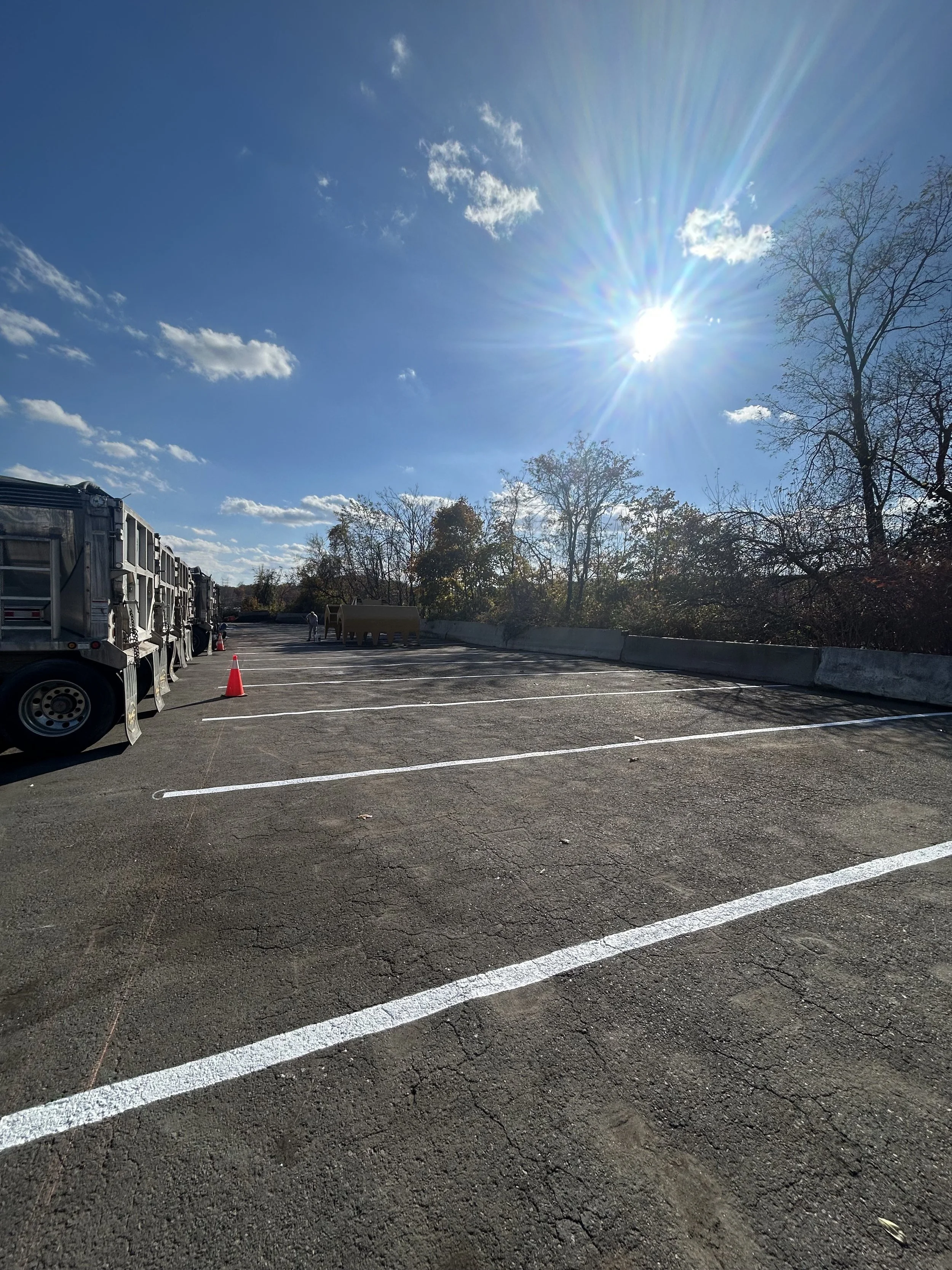 Empty parking lot with a few trucks parked on the left, orange cones, and a bright sun in a clear blue sky with some clouds. Trees with fall foliage are visible in the background.