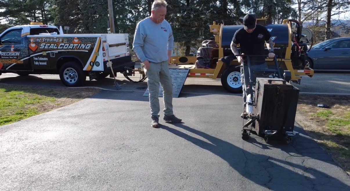 Two men working on a paved driveway, with equipment and trucks in the background.