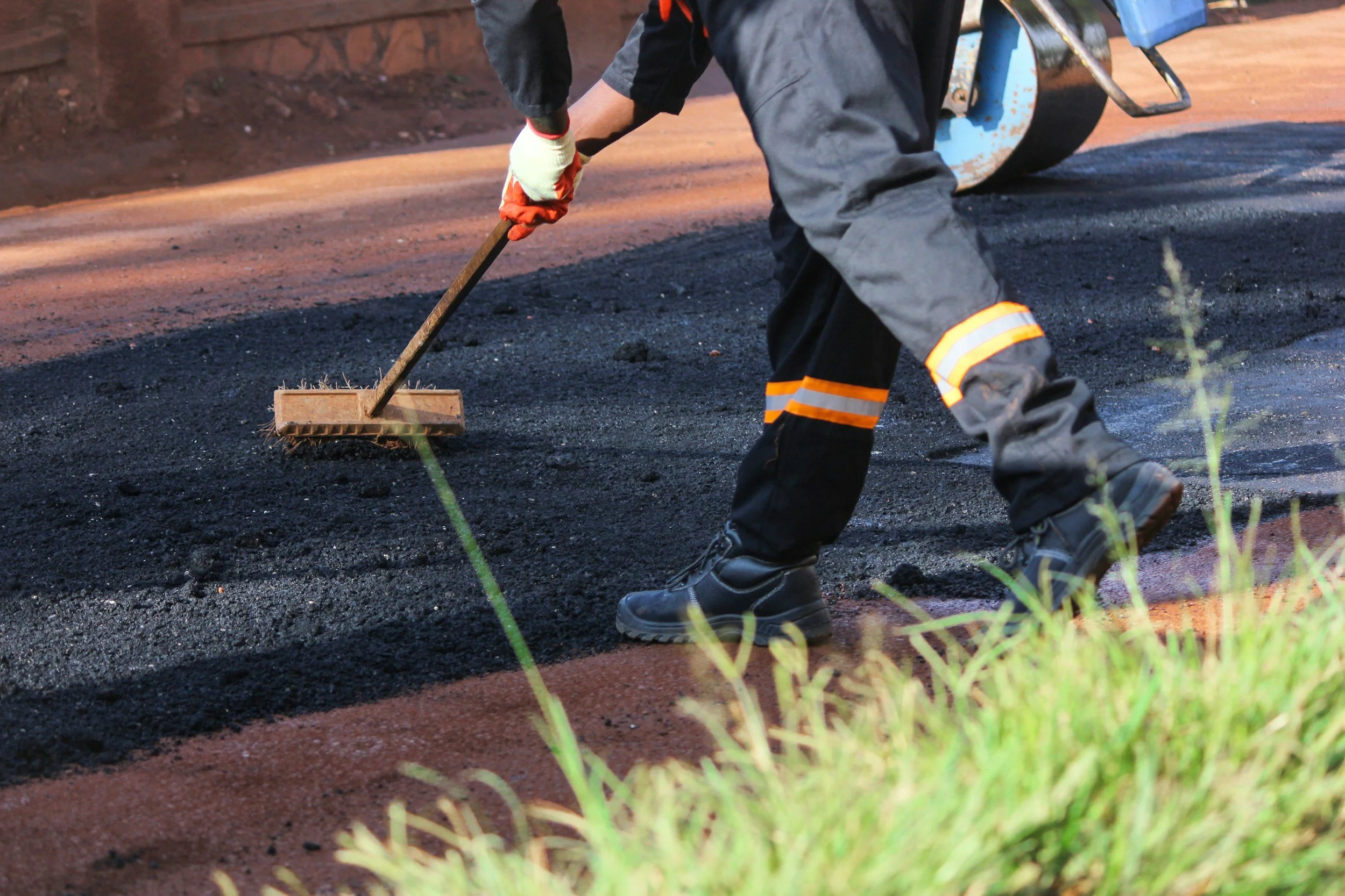 Worker in black safety gear with reflective stripes, wearing gloves and boots, spreads hot asphalt with a rake on a road under construction.