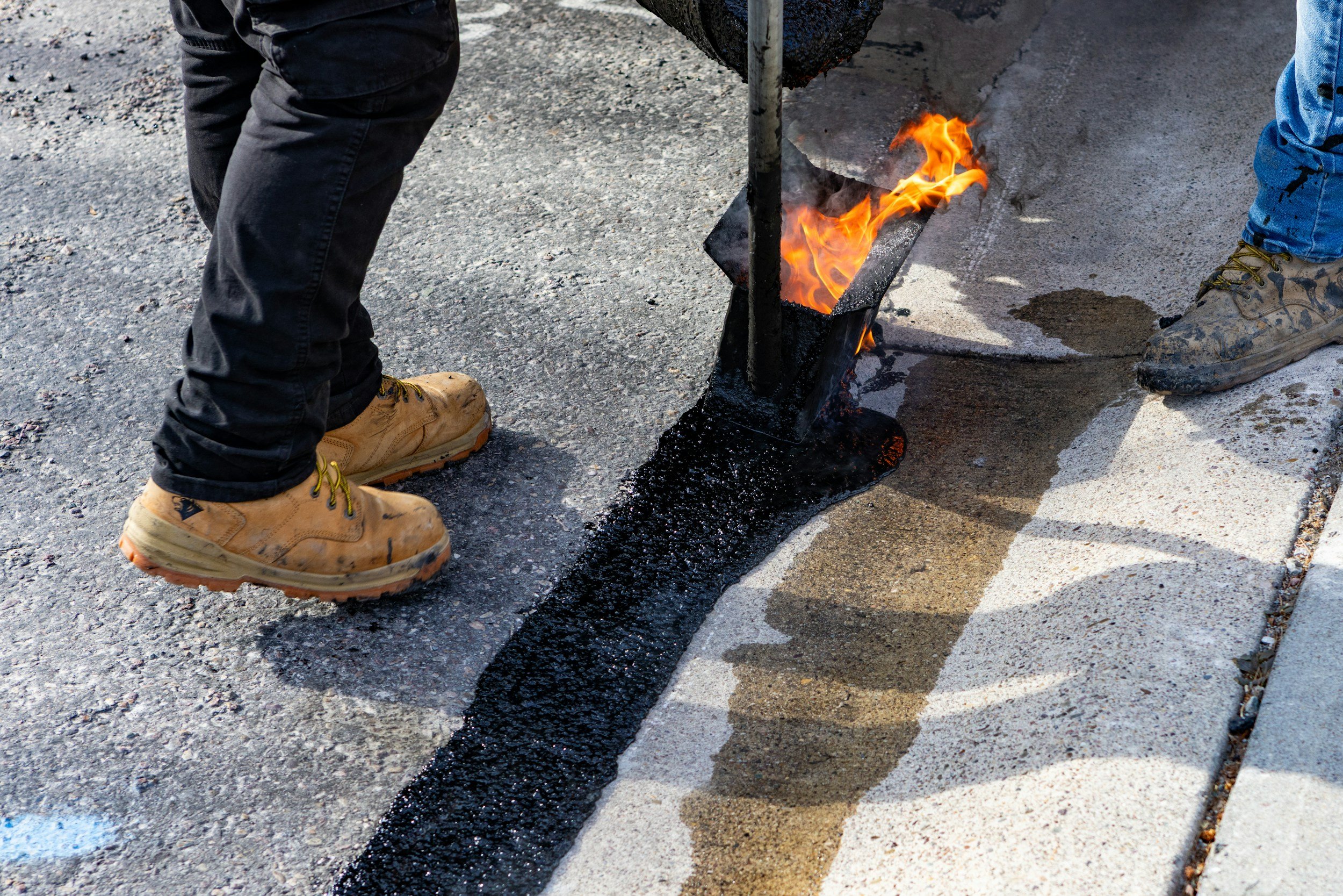 Workers applying hot asphalt to a road, wearing work boots, with a small flame and black asphalt being spread from a tool.