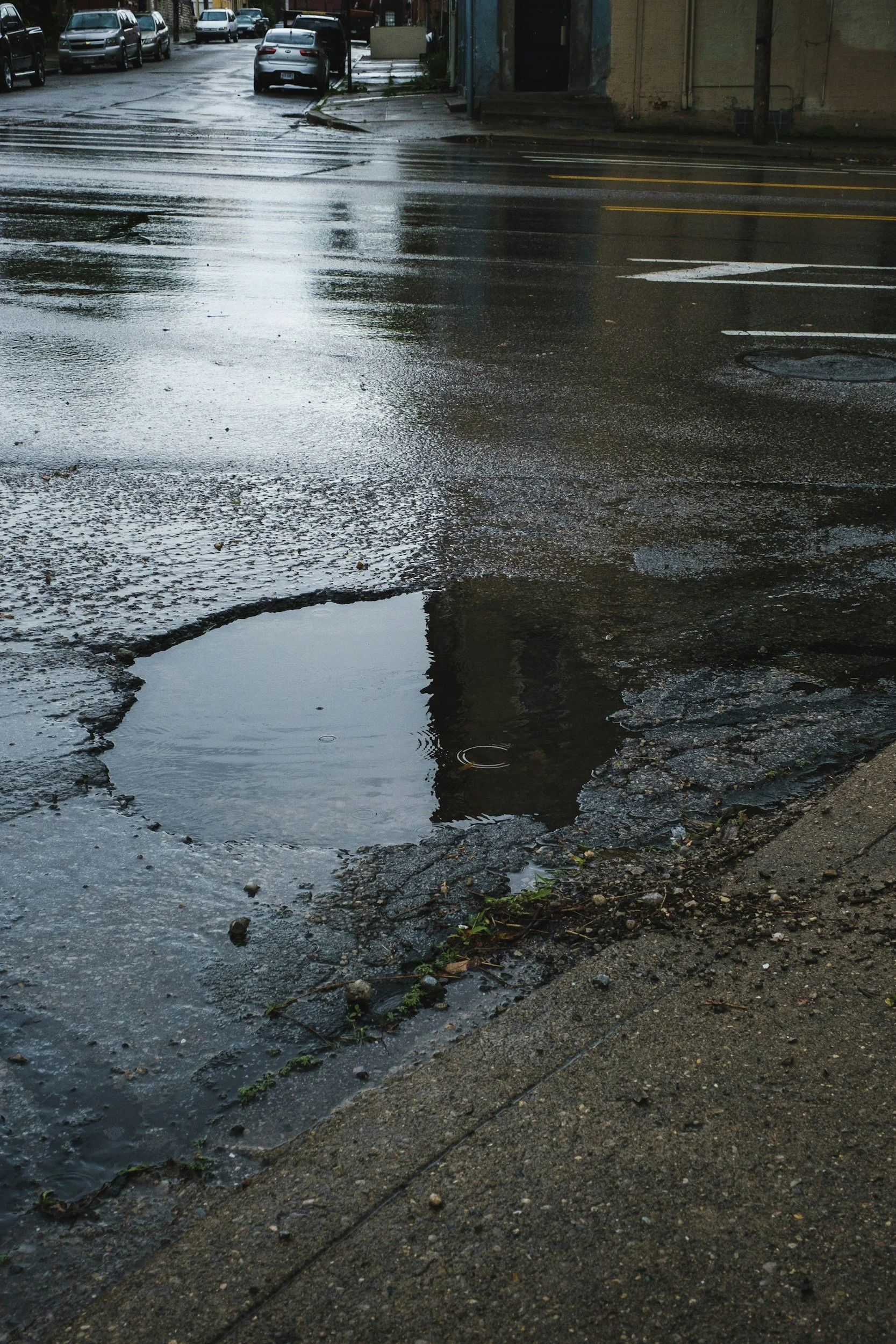 Wet street with large pothole filled with water, located at corner of sidewalk, with parked cars and buildings in background.