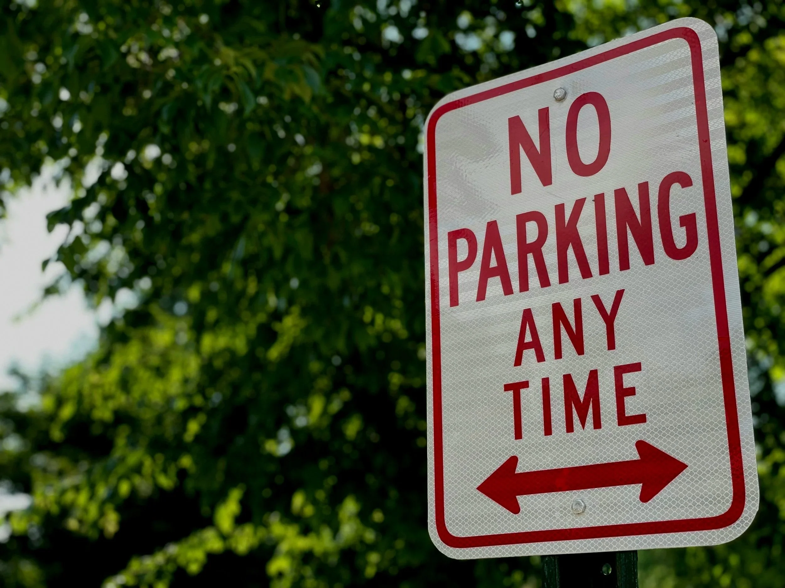 A white and red sign that reads 'NO PARKING ANY TIME' with arrows pointing left and right, against a background of green trees.