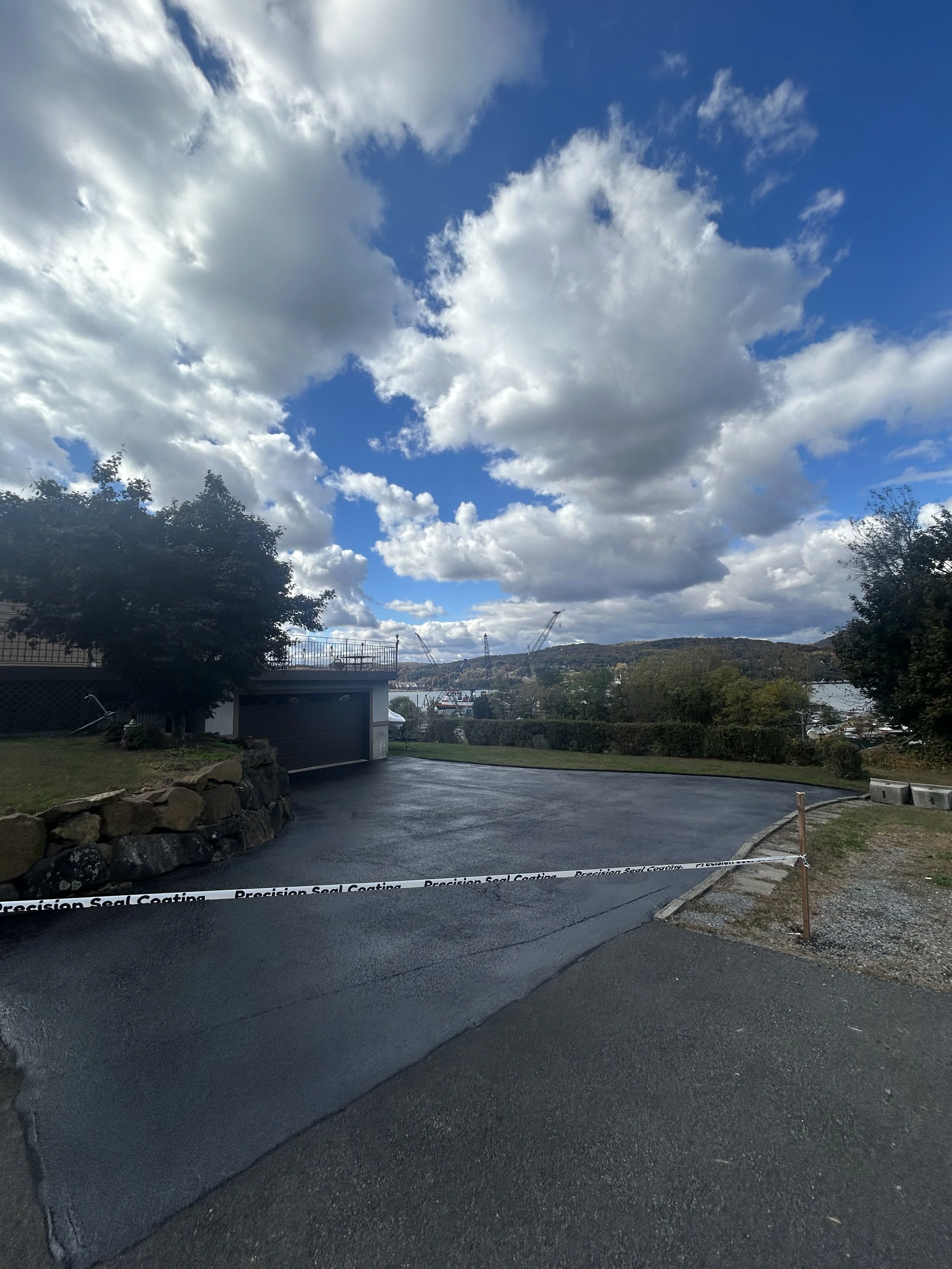 Freshly paved asphalt driveway at a residential property, with a garage and trees, overlooking a body of water under a partly cloudy sky.
