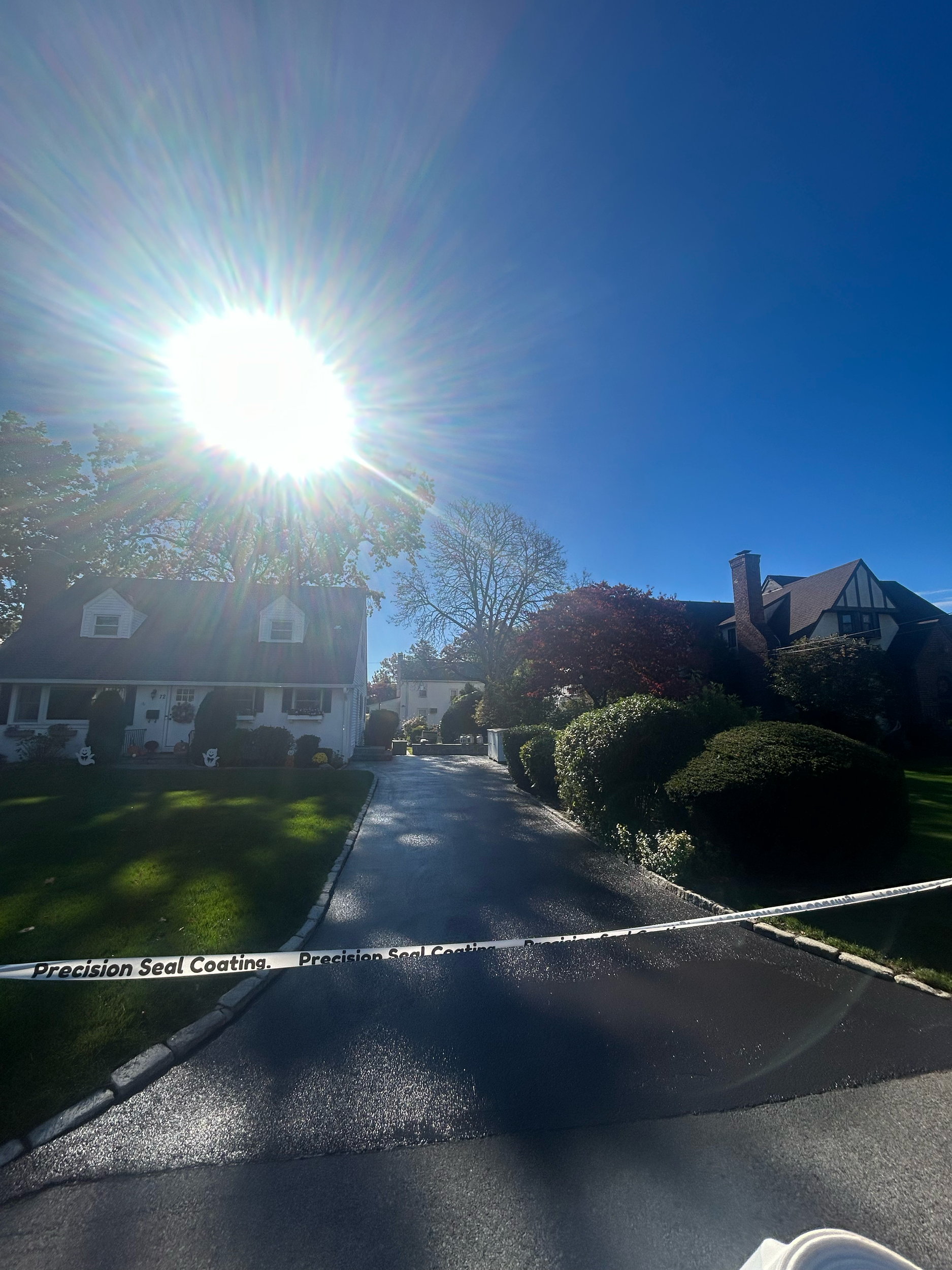 Residential neighborhood street sunny with house, trees, and well-manicured bushes, tar driveway, and caution tape labeled 'Precision Seal Coating' across the foreground.