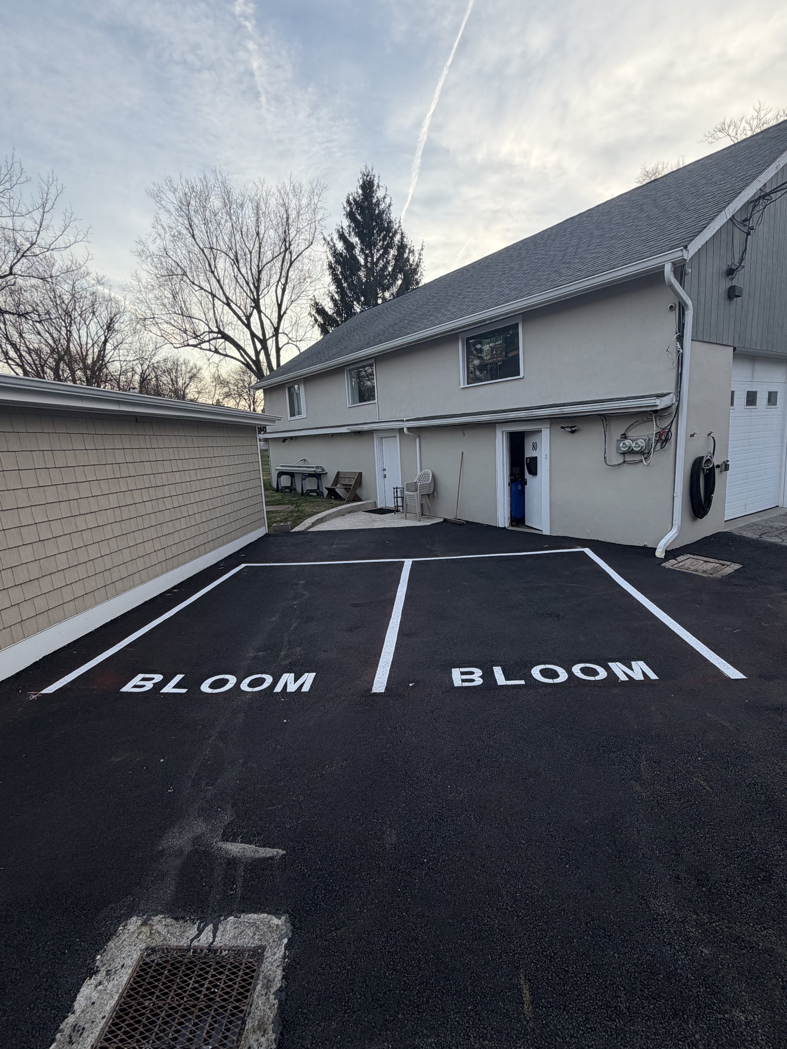 Two parking spaces with white lines and the word 'BLOOM' painted in each space, located in a residential driveway next to a house with a light-colored exterior and a garage door.