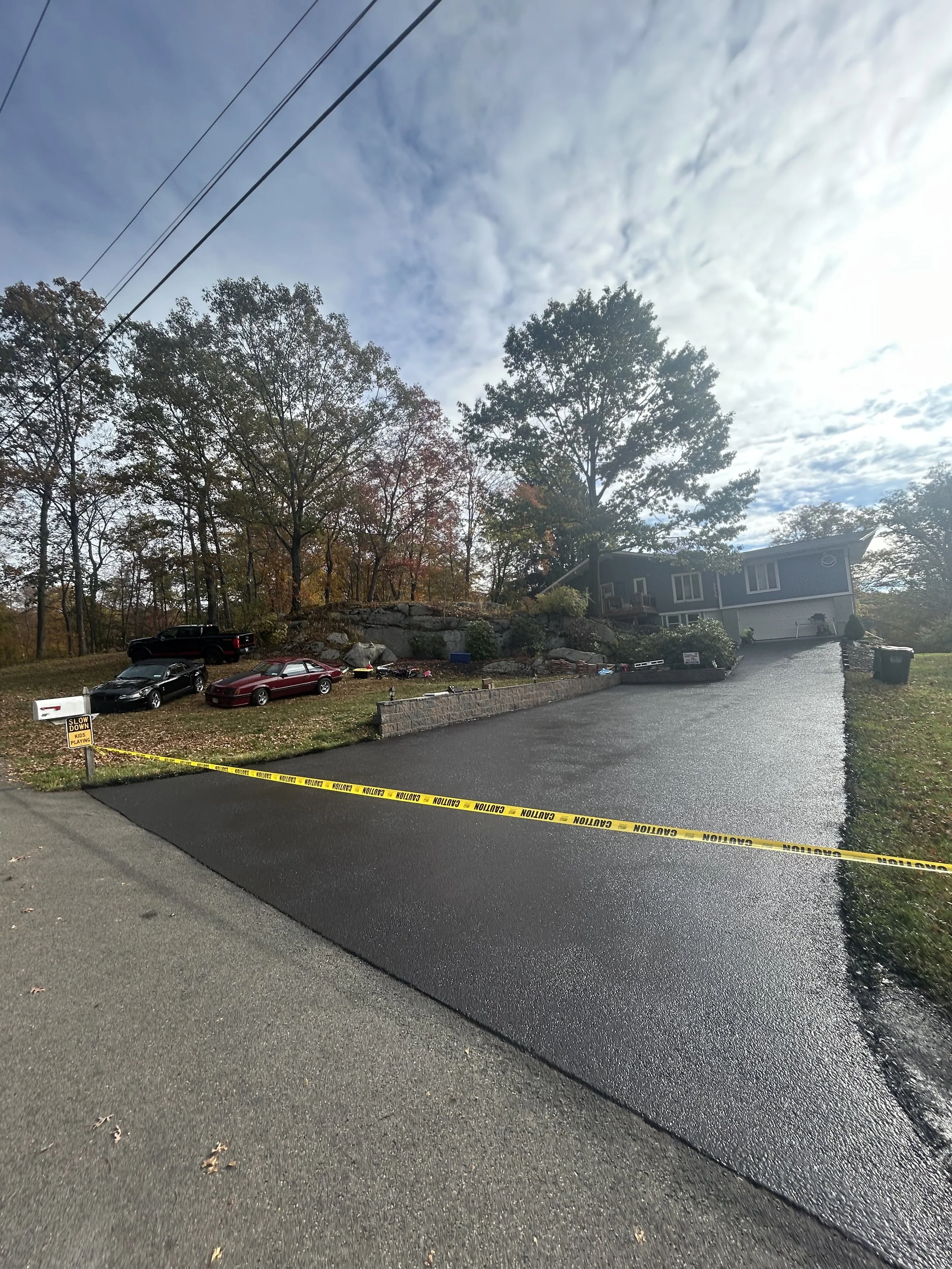 A newly paved black asphalt driveway with a caution tape barrier across the entrance, leading to a house in a suburban area with three parked cars nearby, trees on a hill, and a cloudy sky overhead.