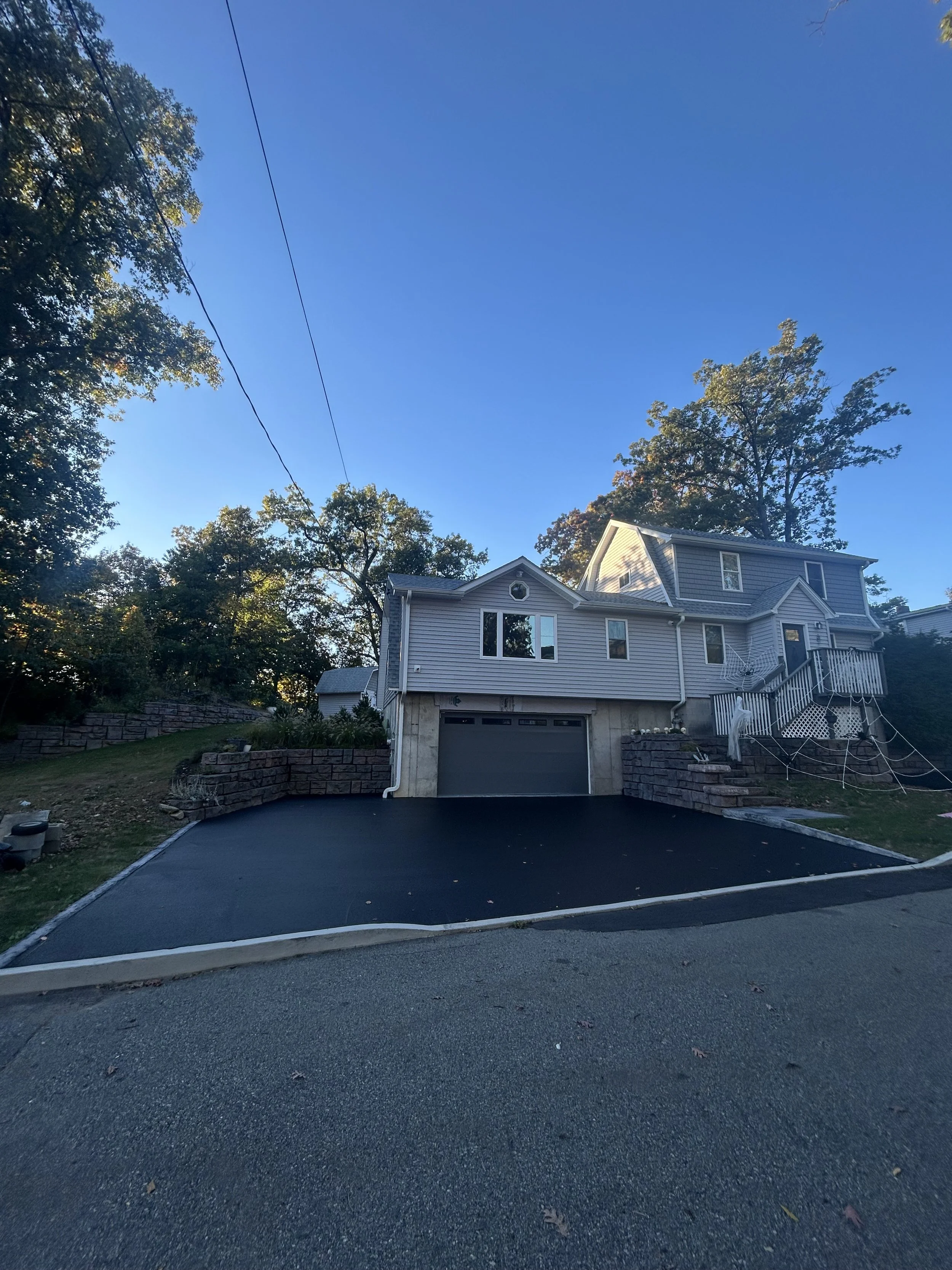 Front view of a house with grey siding, a garage, and a driveway, surrounded by trees and stone retaining walls, under a clear blue sky.