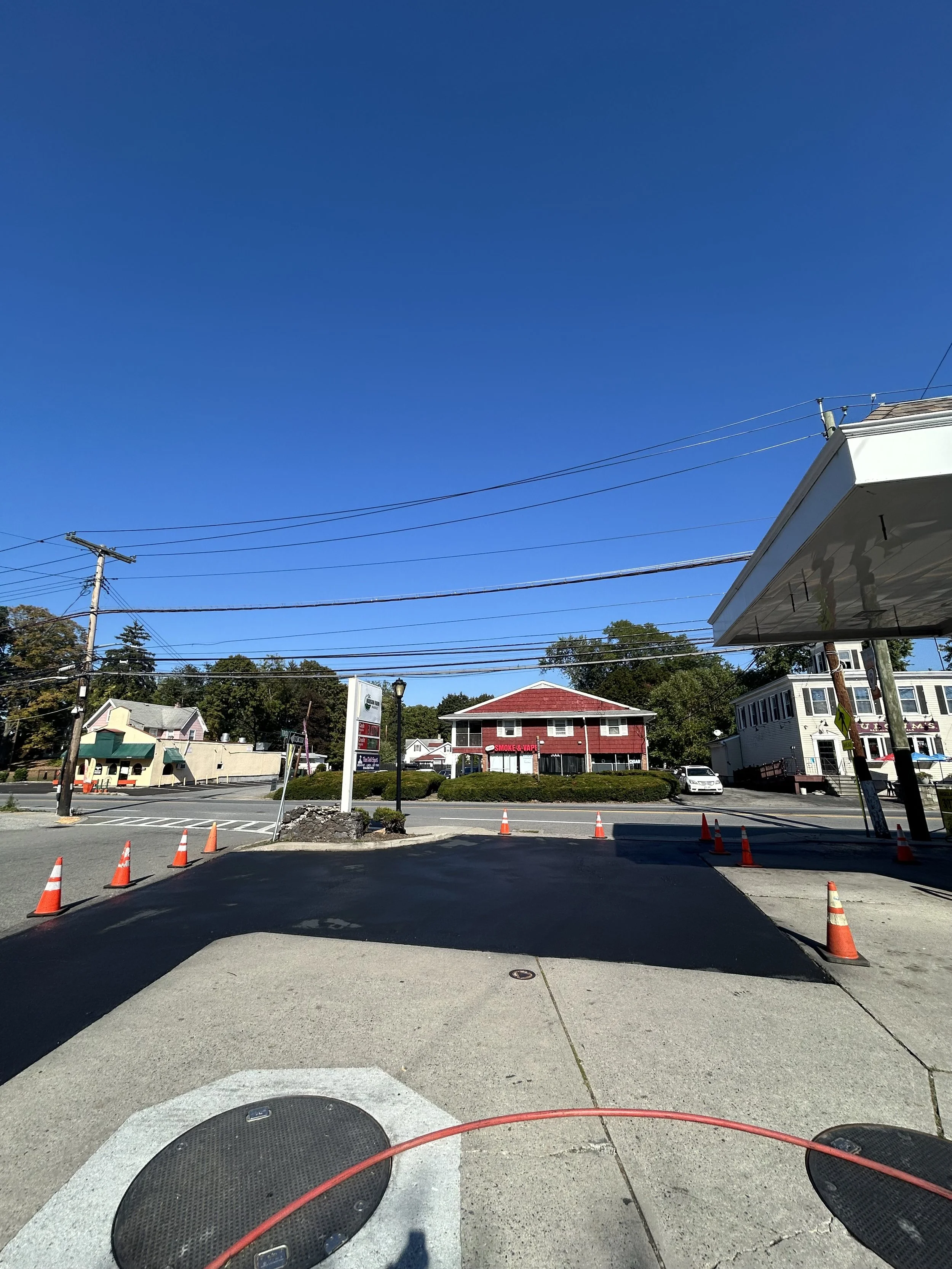 A sunny street view with a blue sky, utility poles, and wires. There are buildings, including a red building with a sign and a white building, along with orange traffic cones in the foreground.