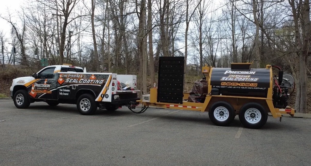A white pickup truck connected to a yellow trailer with a black tank, parked in a lot with leafless trees in the background. Both vehicles display branding for a seal coating company.