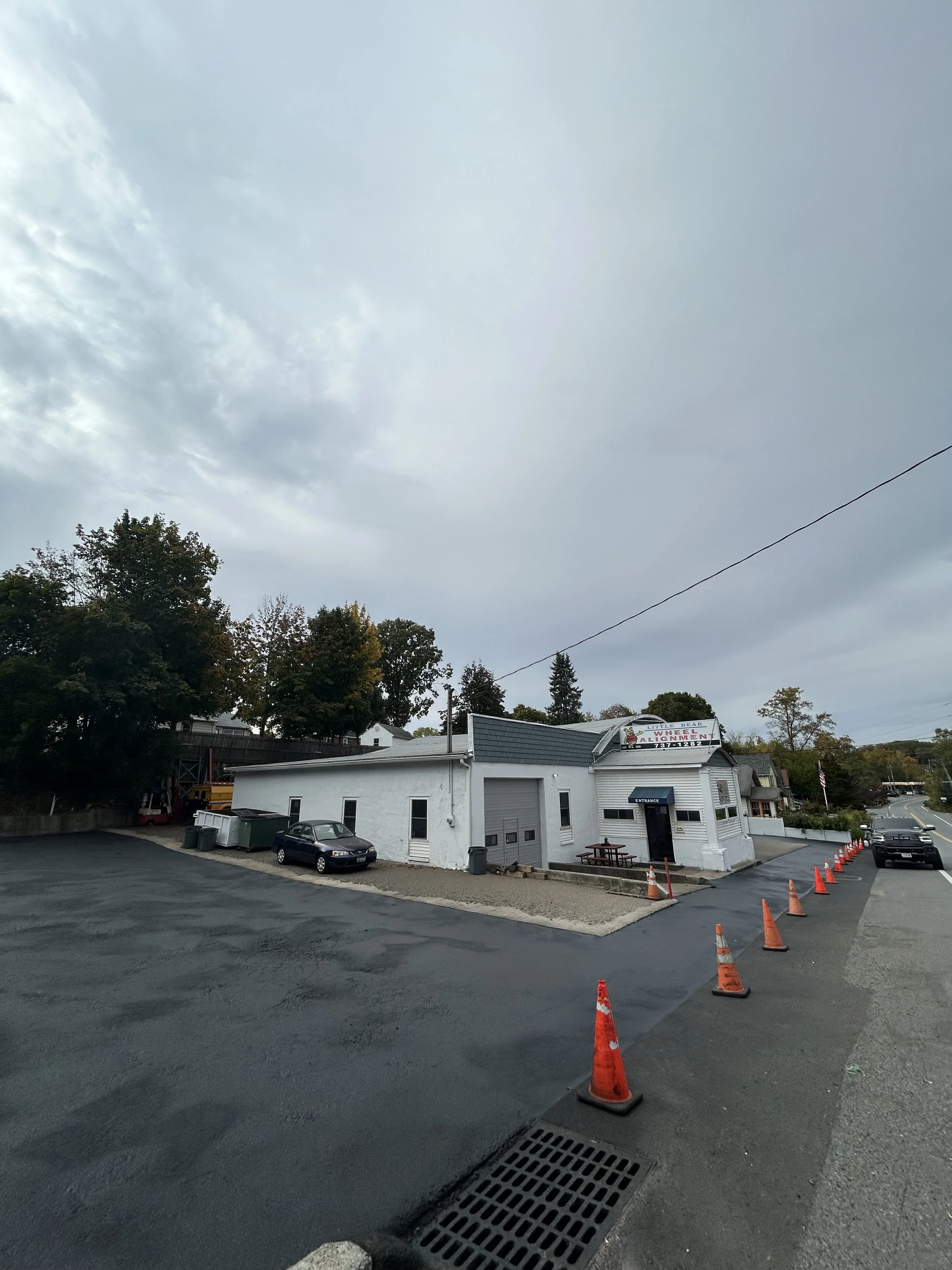 A white building with a parking lot in front, parking cones, and a sign for a business. Parked cars are visible nearby, and trees and houses are in the background under a cloudy sky.