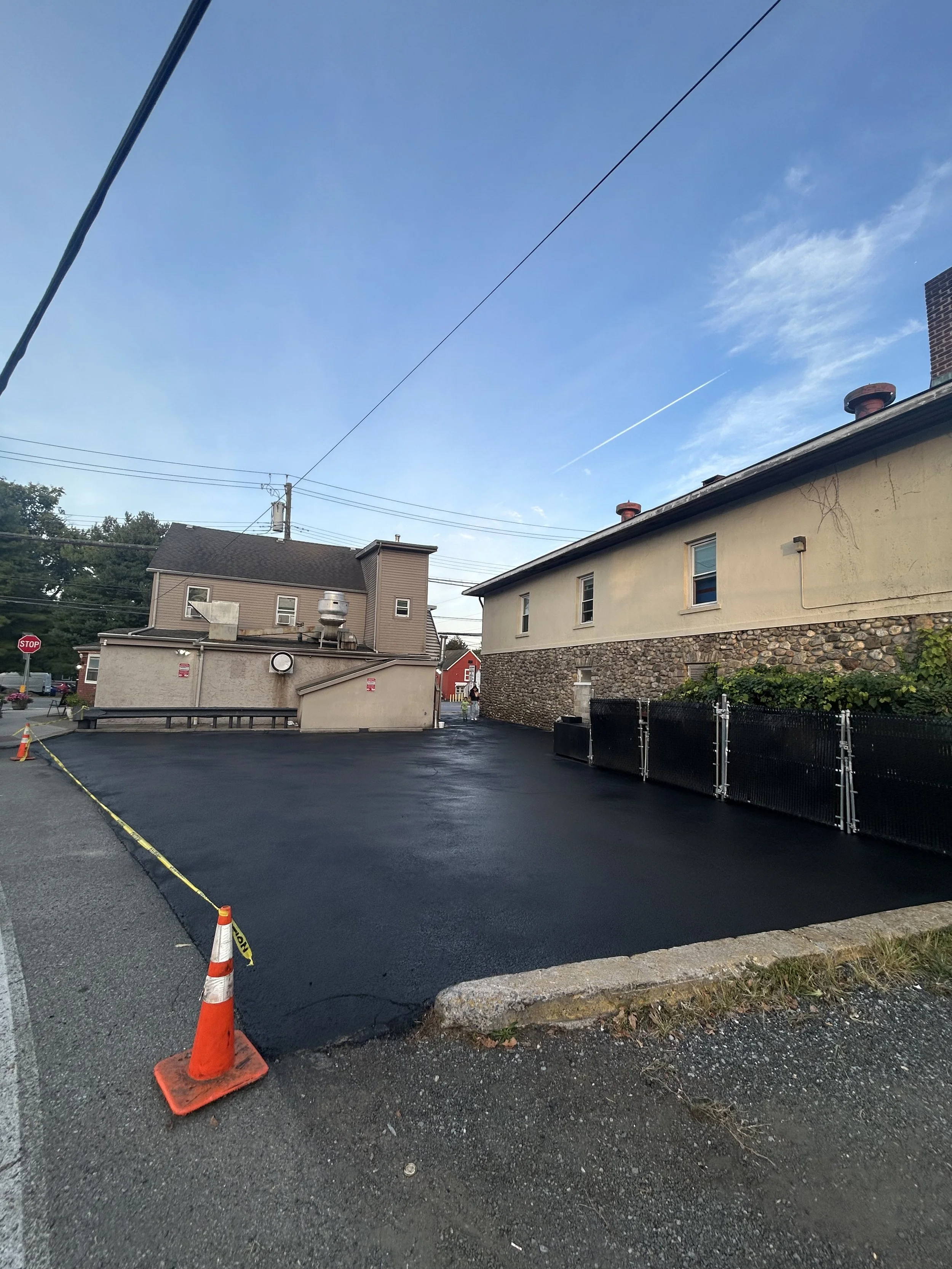 Freshly paved asphalt lot next to residential buildings, orange traffic cone and caution tape in foreground, blue sky with a few clouds overhead.