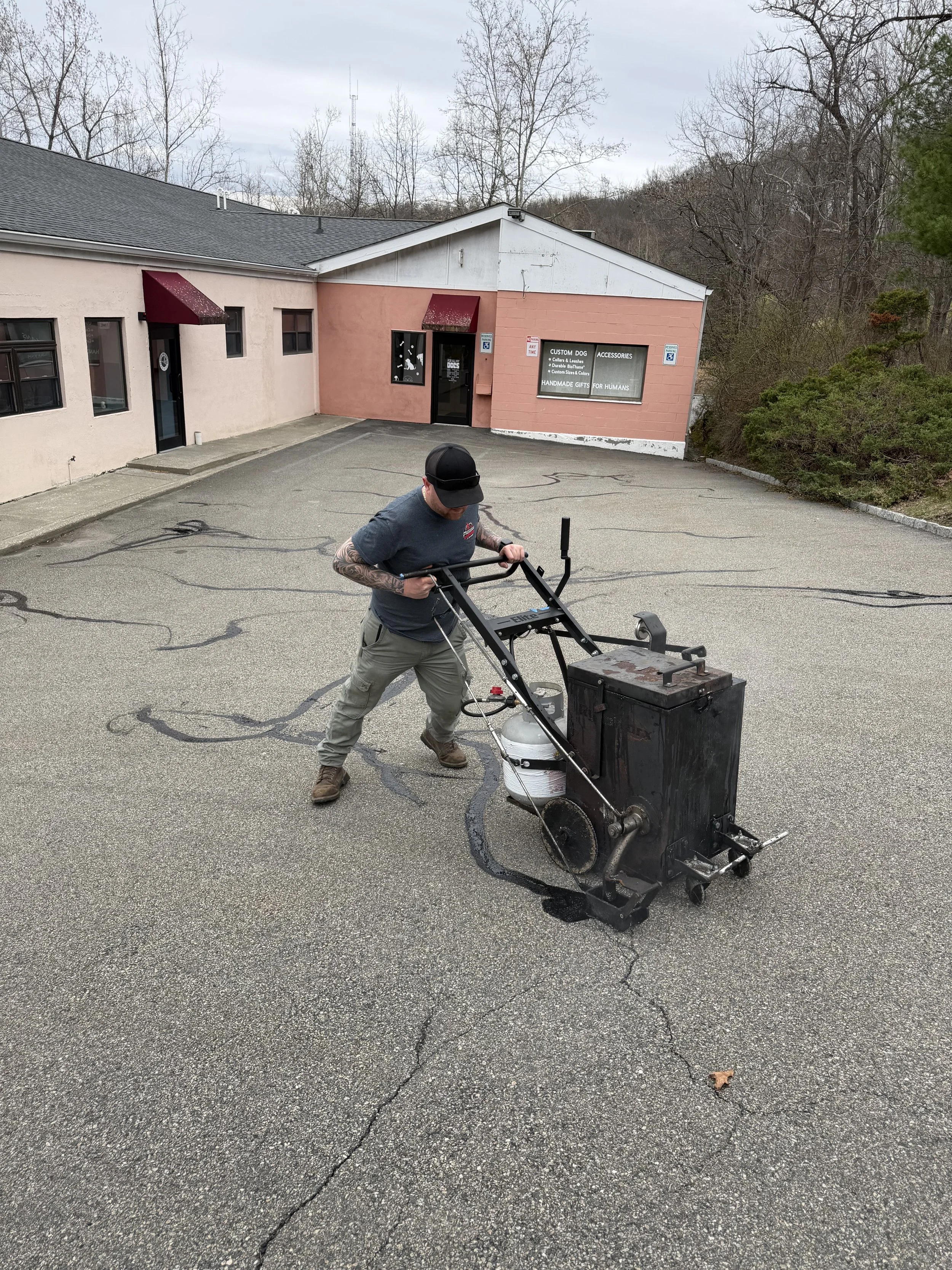 A man operating a crack sealing machine on an asphalt parking lot outside a pink building with signs for a custom dog shop.
