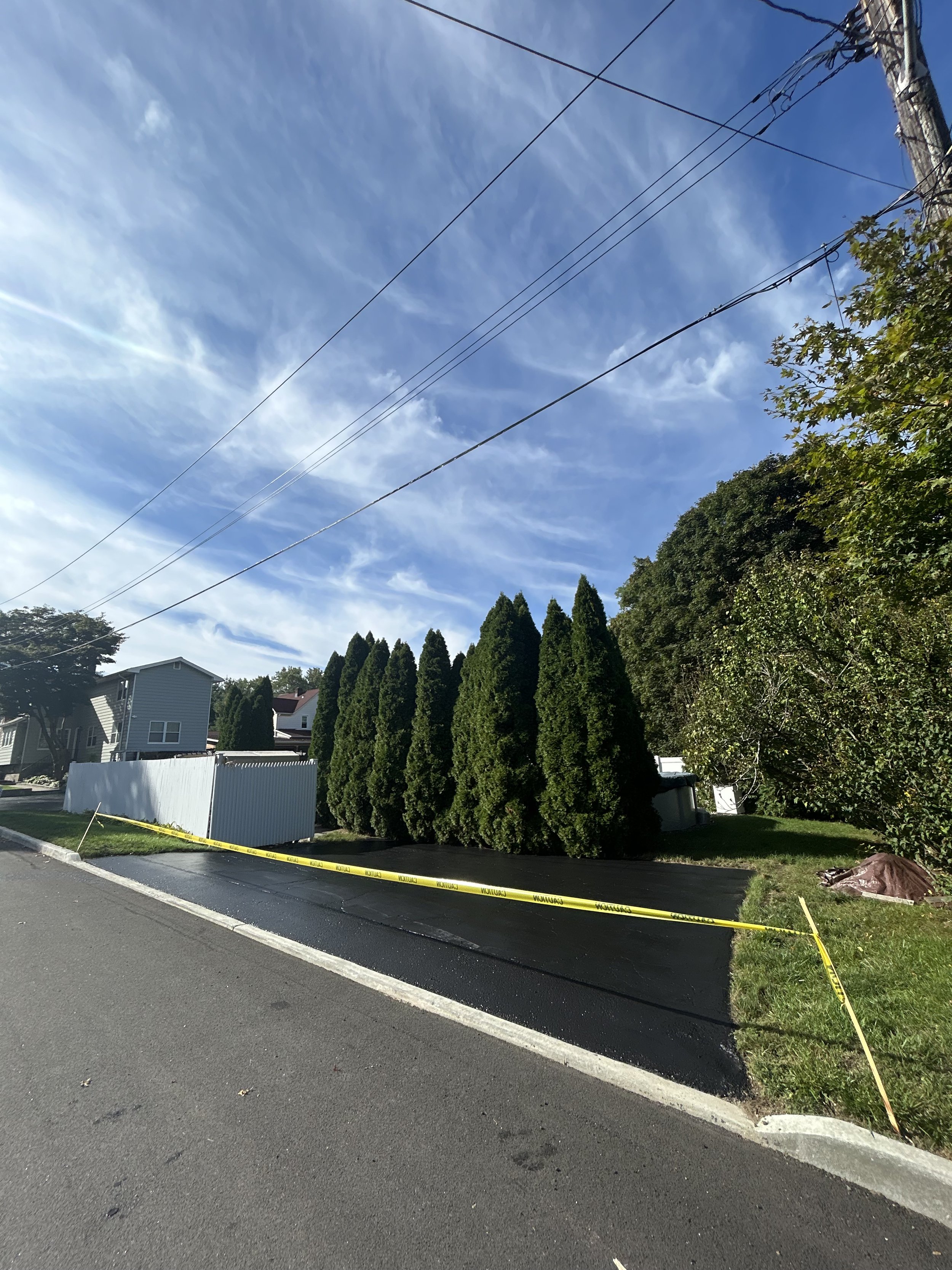 A residential street with a freshly paved driveway, yellow caution tape across, and tall green trees and bushes along the sidewalk. Power lines extend across the bright blue sky with wispy clouds.
