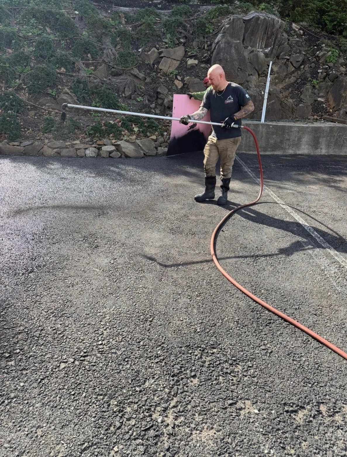 Person wearing black t-shirt, khaki pants, and rubber boots smoothing freshly laid asphalt on a road with a hose, with another person in the background involved in the work.