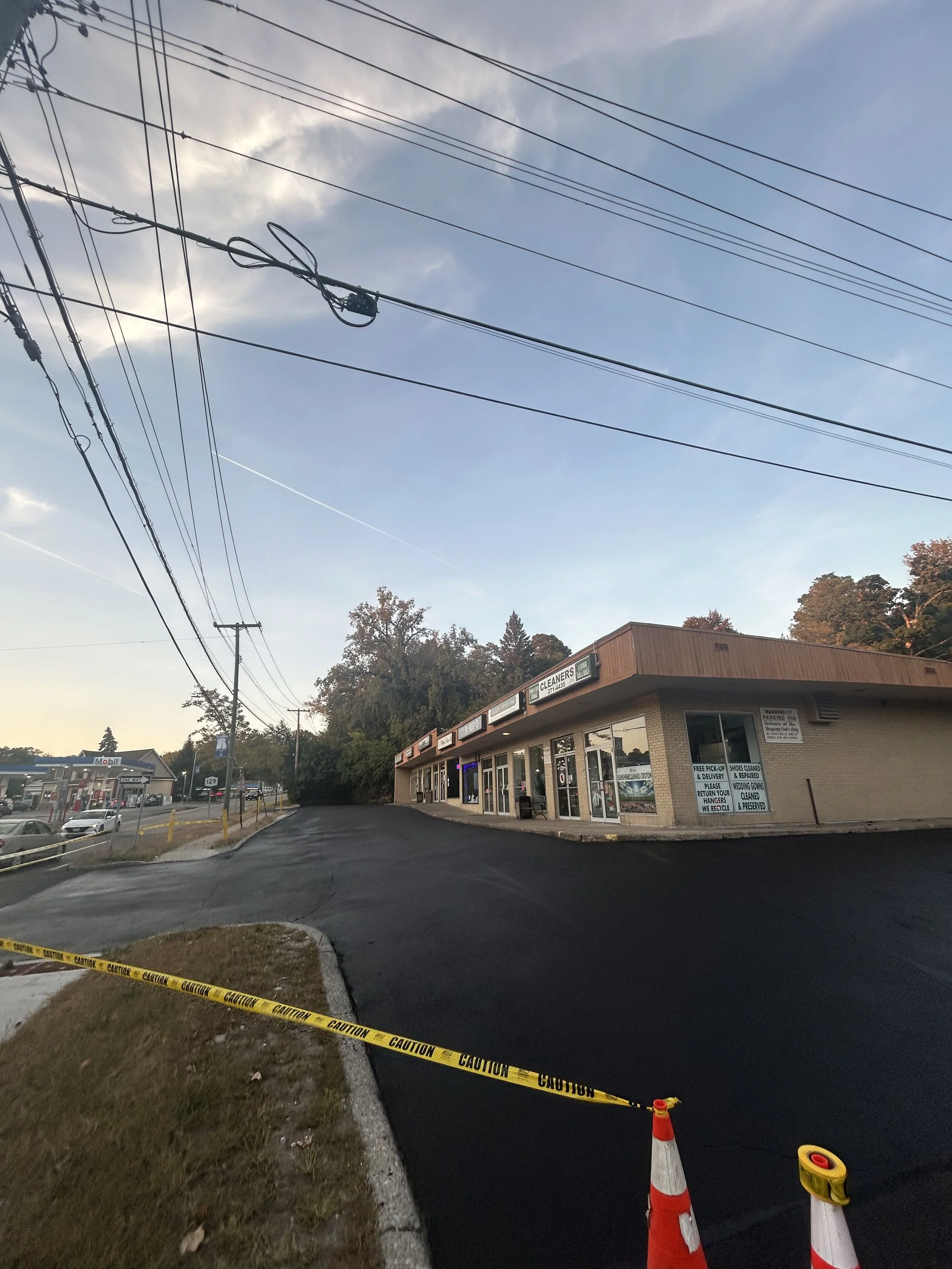 Newly paved parking lot in front of a small commercial building with a sign for a cleaning business, caution tape and traffic cones in the foreground, power lines overhead, and a gas station in the background on the left.