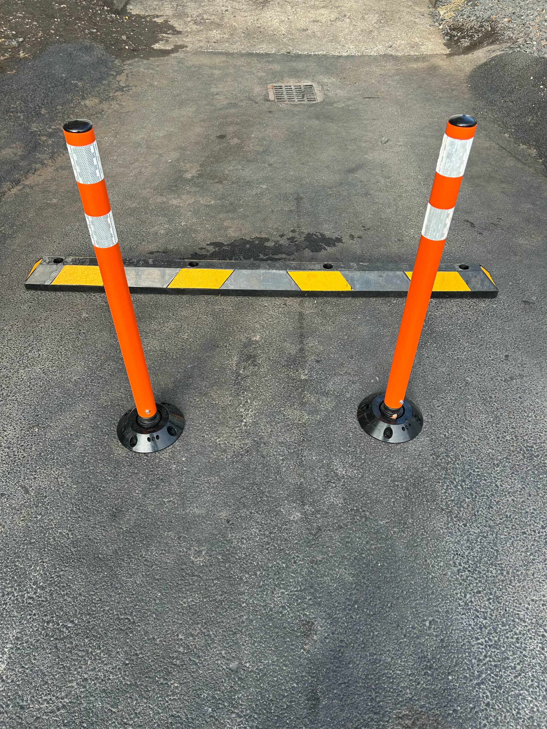Two orange traffic posts with reflective strips and black bases are positioned in front of a parking space barrier on a paved surface.