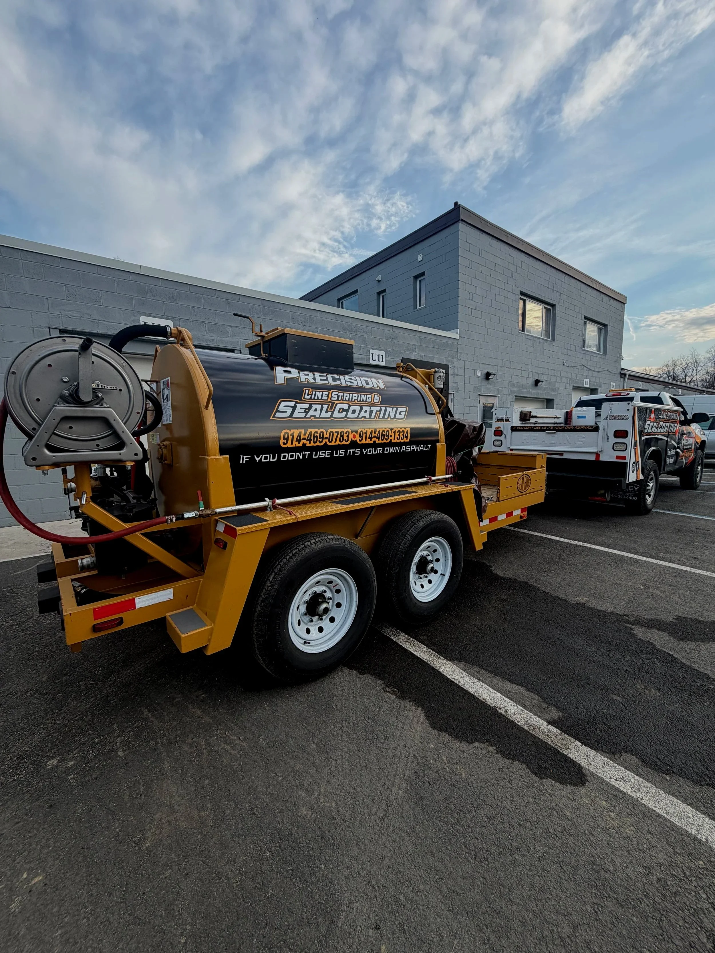 A yellow trailer with a black tank for sealing and coating services parked in a lot, with a gray building in the background and a partly cloudy sky above.