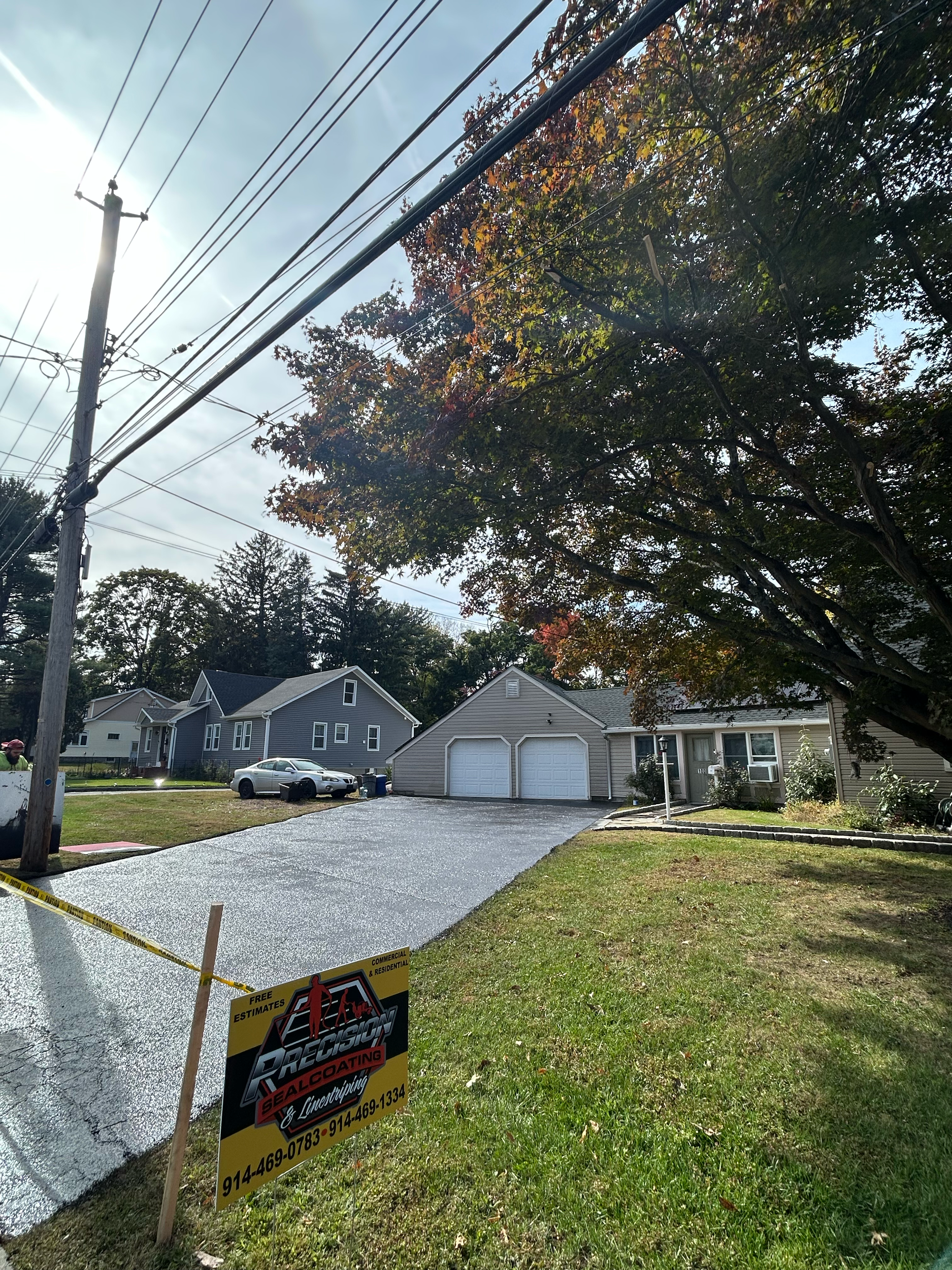 Residential area with freshly sealed driveway, three houses in background, large tree to the right, utility pole and power lines above, sign in front advertising sealing services with contact information.