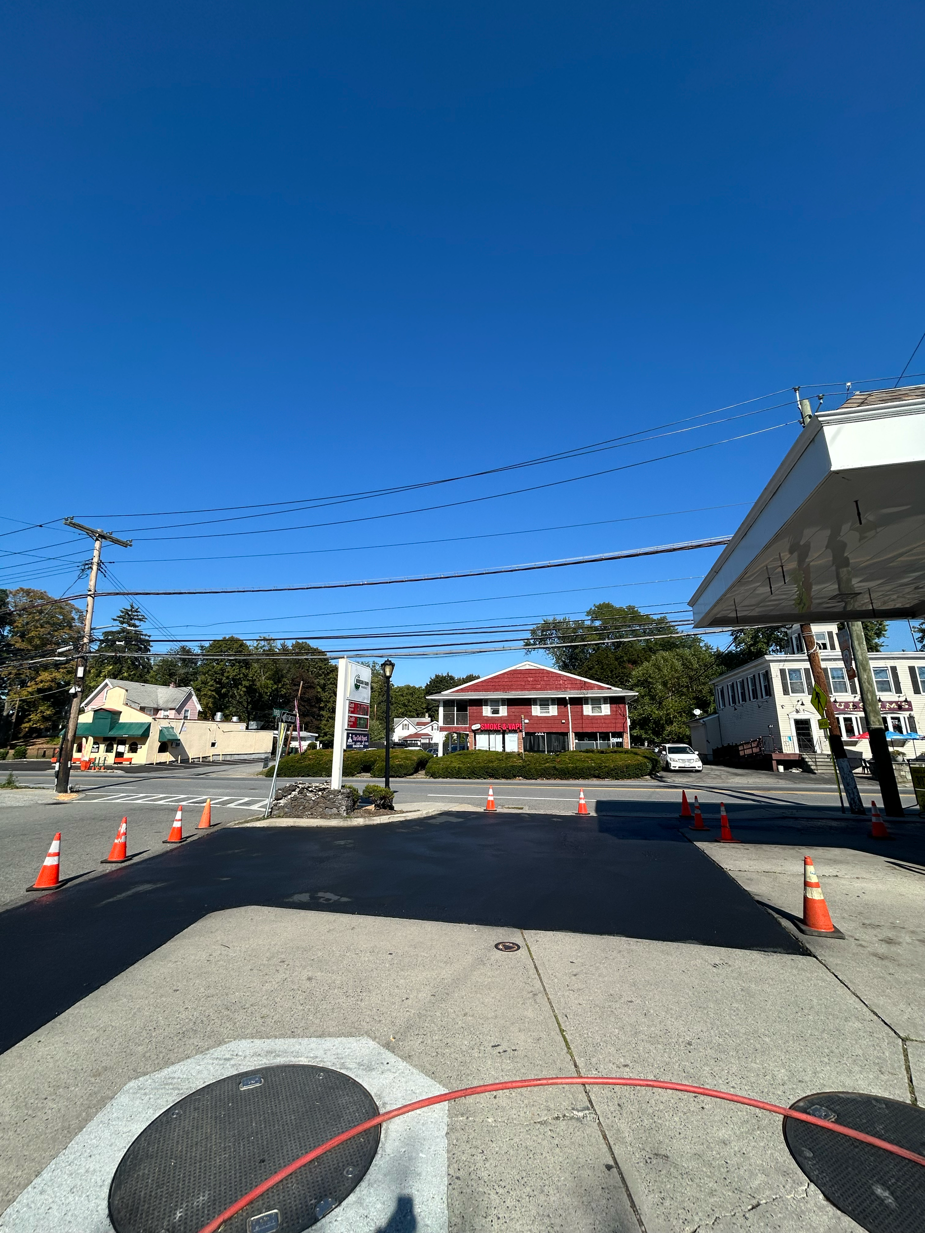 A street view with a clear blue sky, traffic cones blocking part of the road, and buildings including a red house, a white building, and a green storefront.
