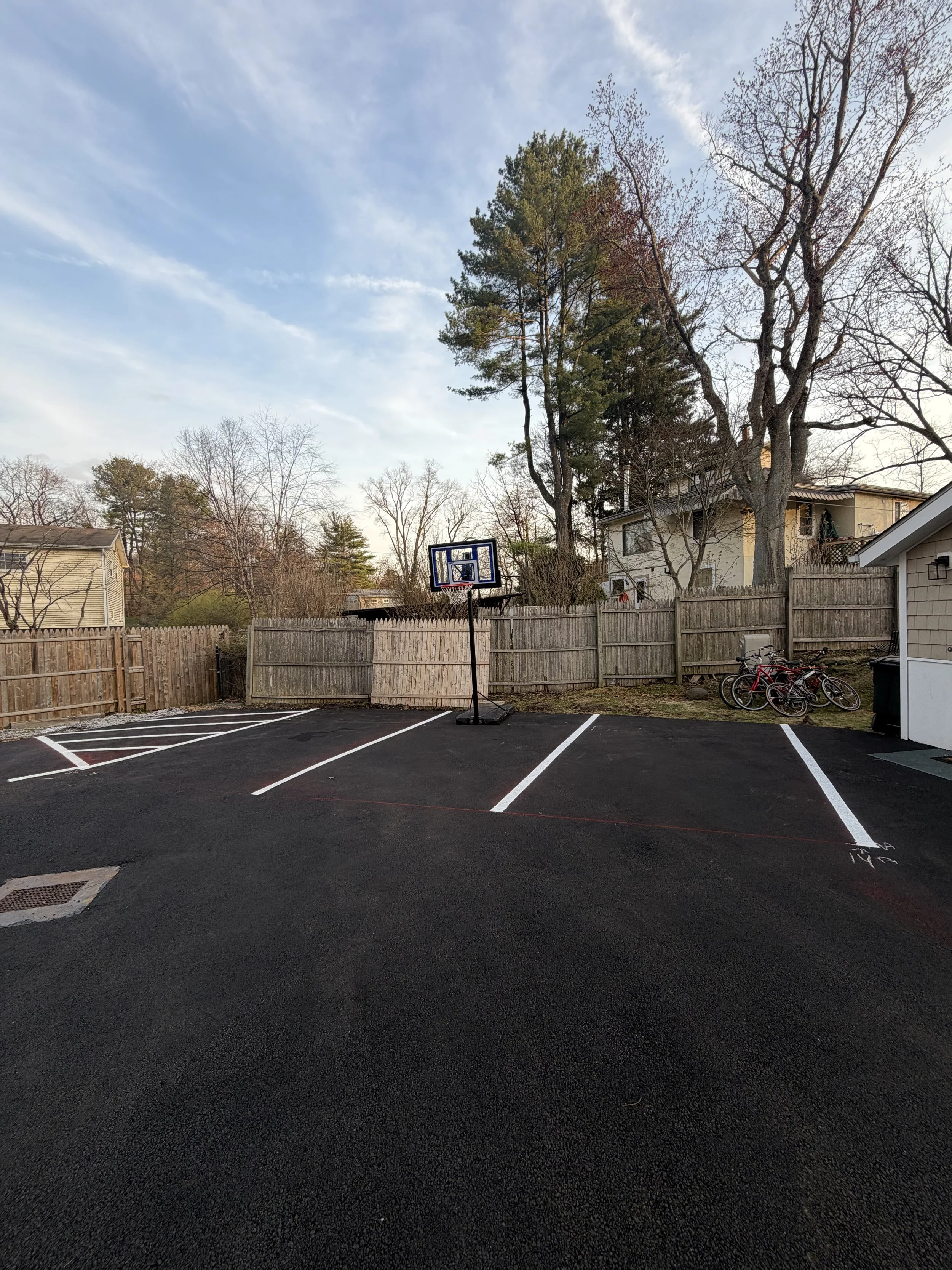 A small outdoor sports court with a basketball hoop, empty parking spaces, and a wooden fence, surrounded by trees and residential houses.