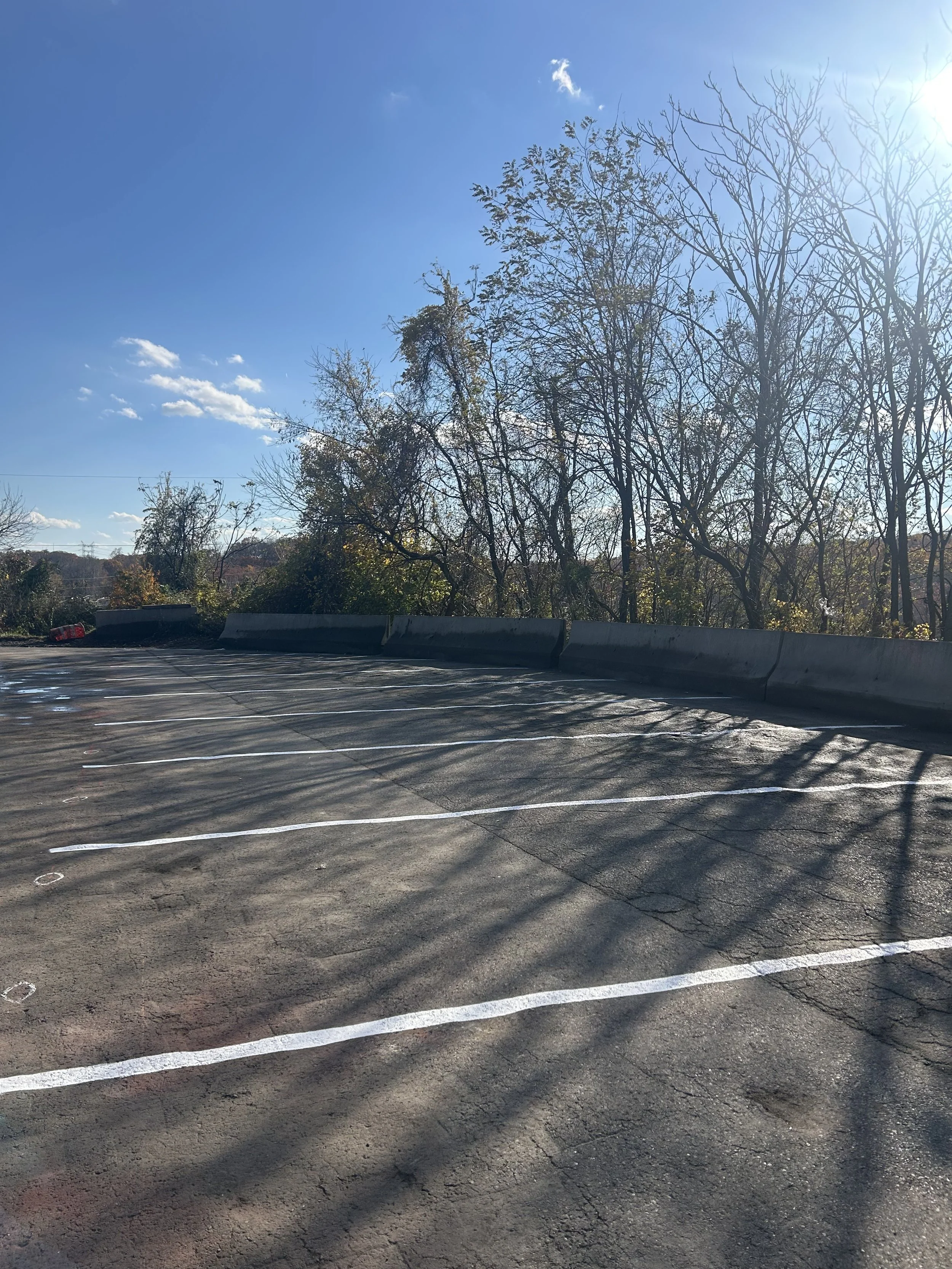 Empty parking lot with white parking lines on asphalt, concrete barriers at the edge, trees without leaves, and a clear blue sky with some clouds.
