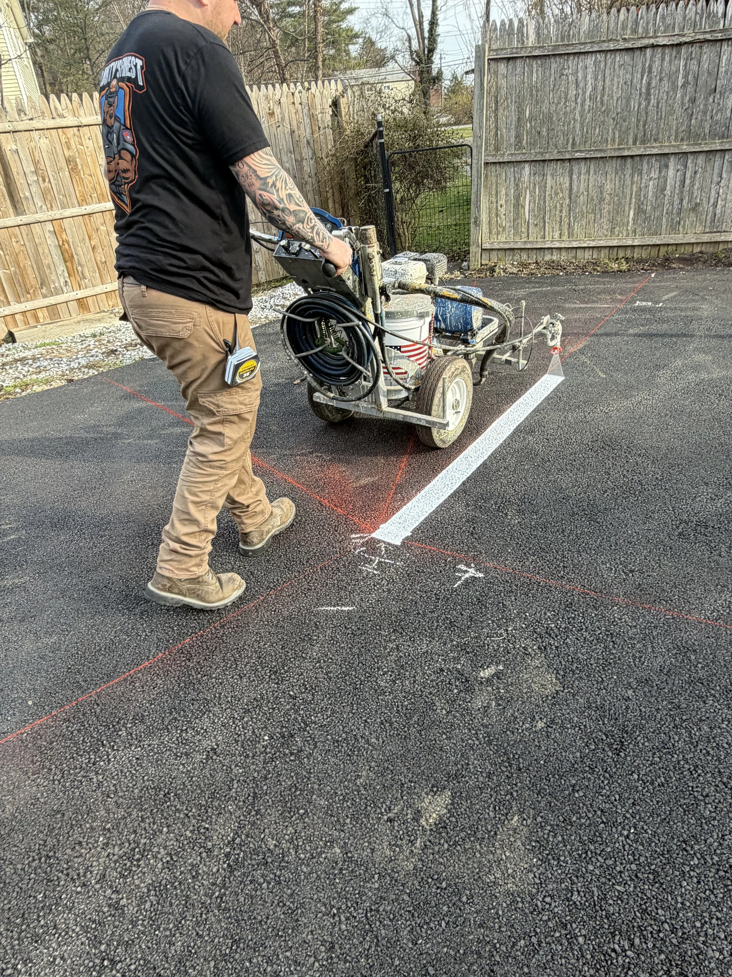 A worker is preparing to paint parking space lines with a line striping machine on a paved surface, with chalk markings and red spray lines visible on the ground.