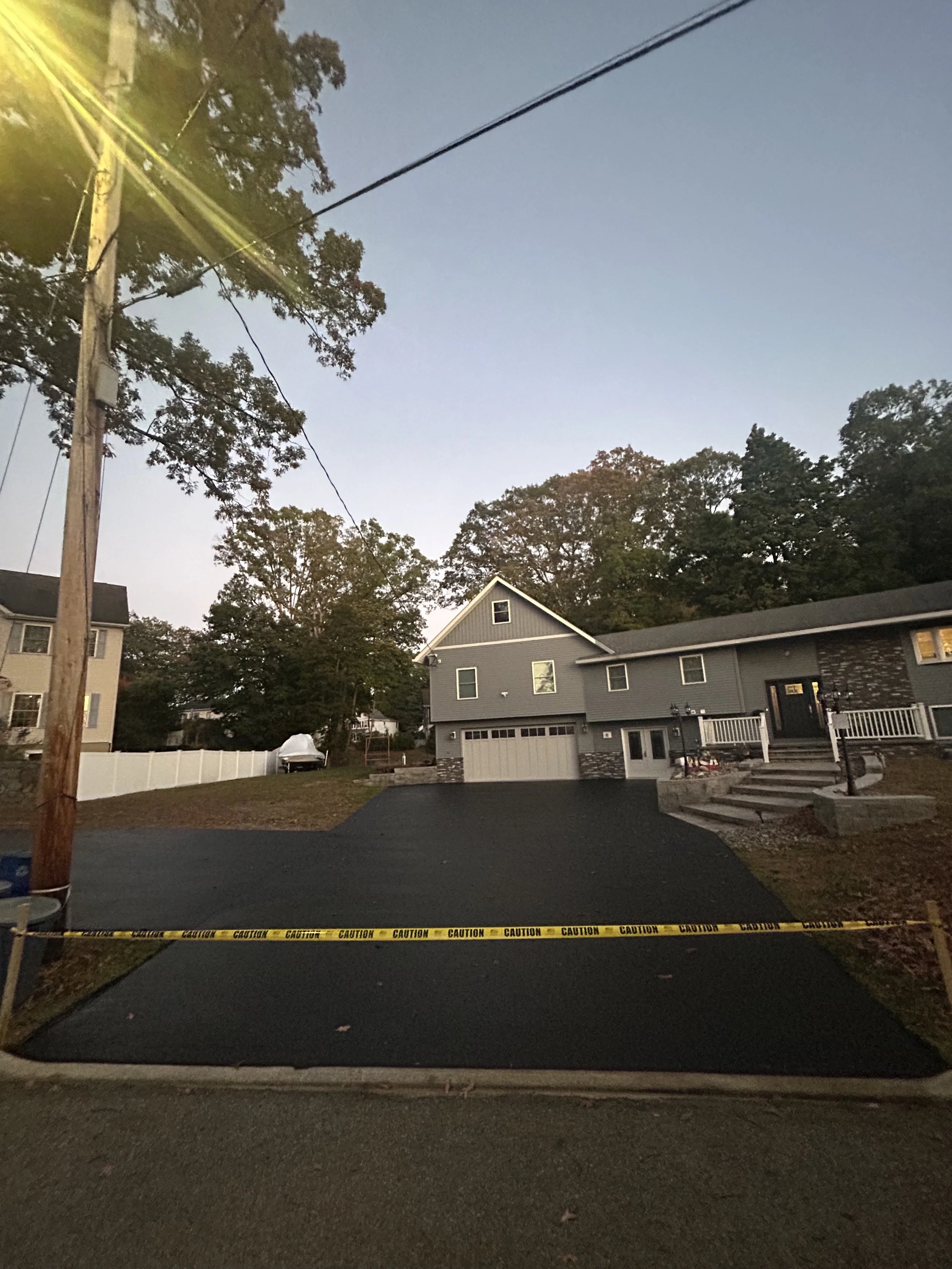 Newly paved driveway with caution tape, leading to a two-story gray house with white trim and front porch, surrounded by trees, under a dusk sky.