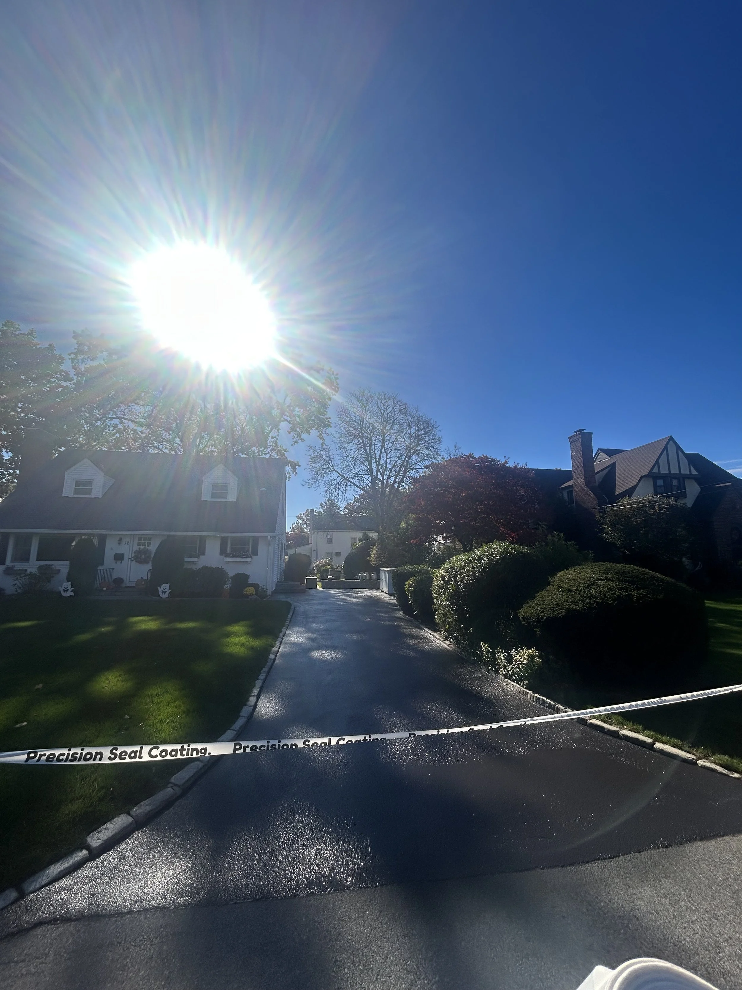 Sunny day over a suburban residential street with houses, trees, and shrubs, with a section of caution tape reading 'Precision Seal Coating' crossing the driveway.