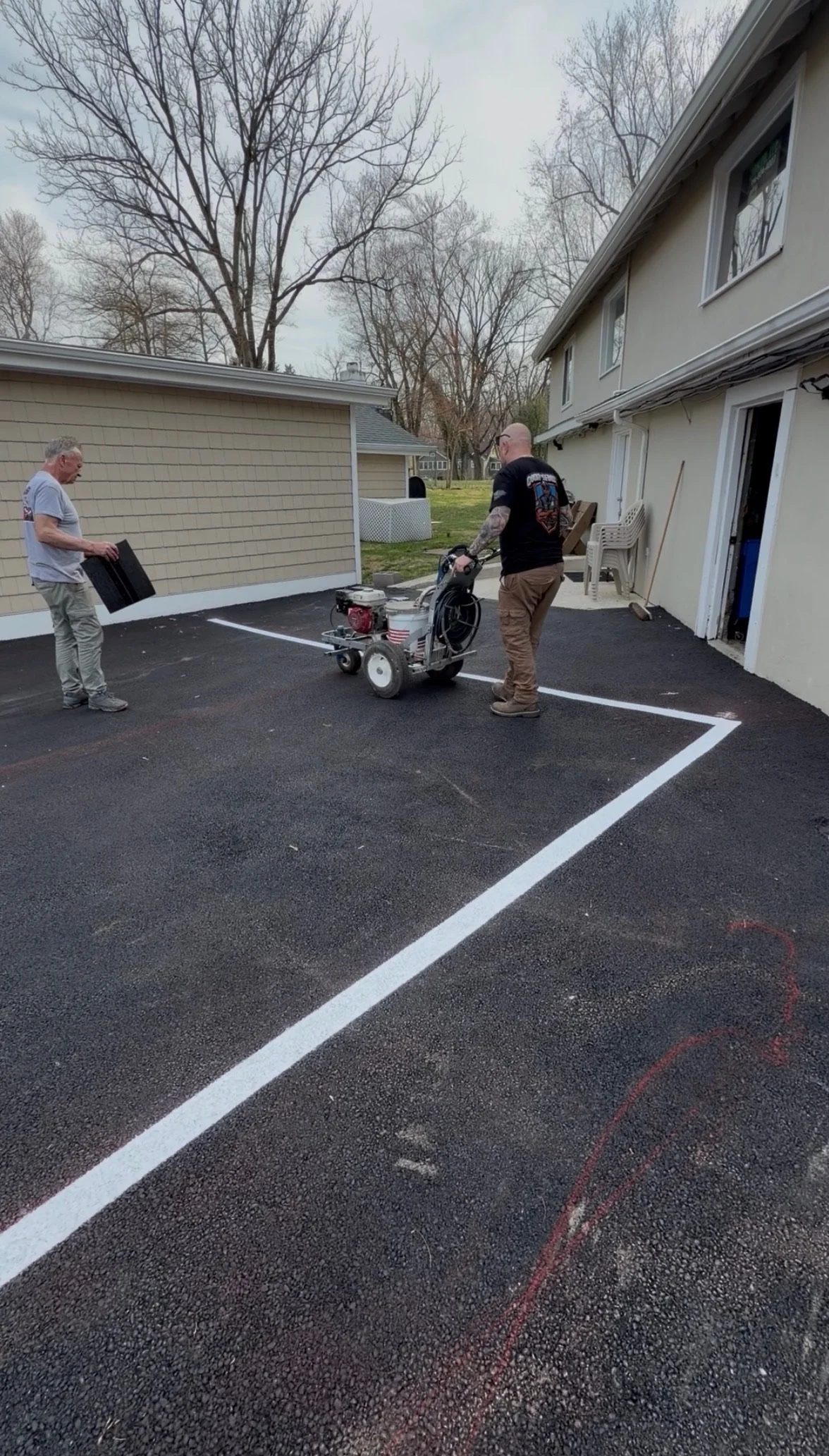 Two men working on parking lot line painting outside a house. One man is holding a paint tray, while the other operates line marking equipment.