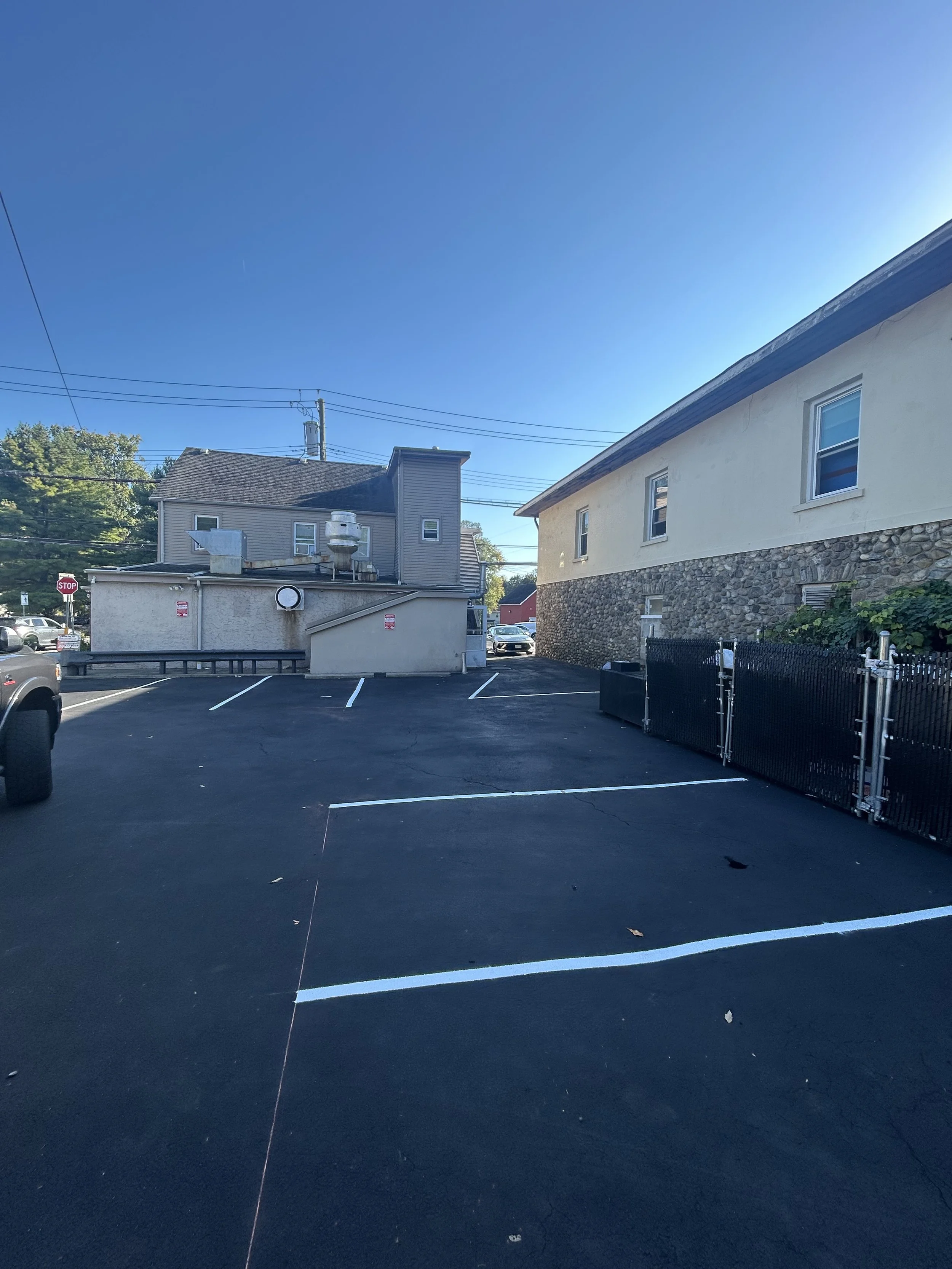 Empty parking lot with white parking lines, black asphalt, and two gated entrances, beside a beige building with stone foundation, against clear blue sky.