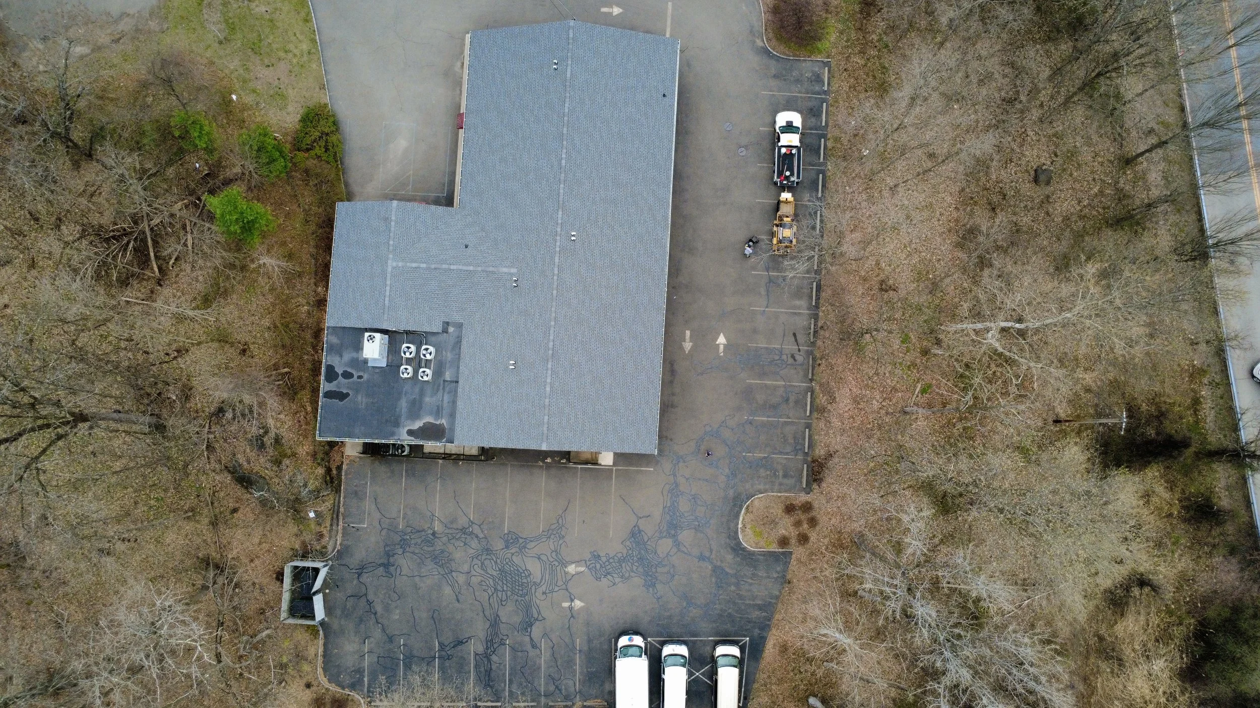 Aerial view of a building with a parking lot surrounded by trees, some parked cars, and utility equipment on the roof.