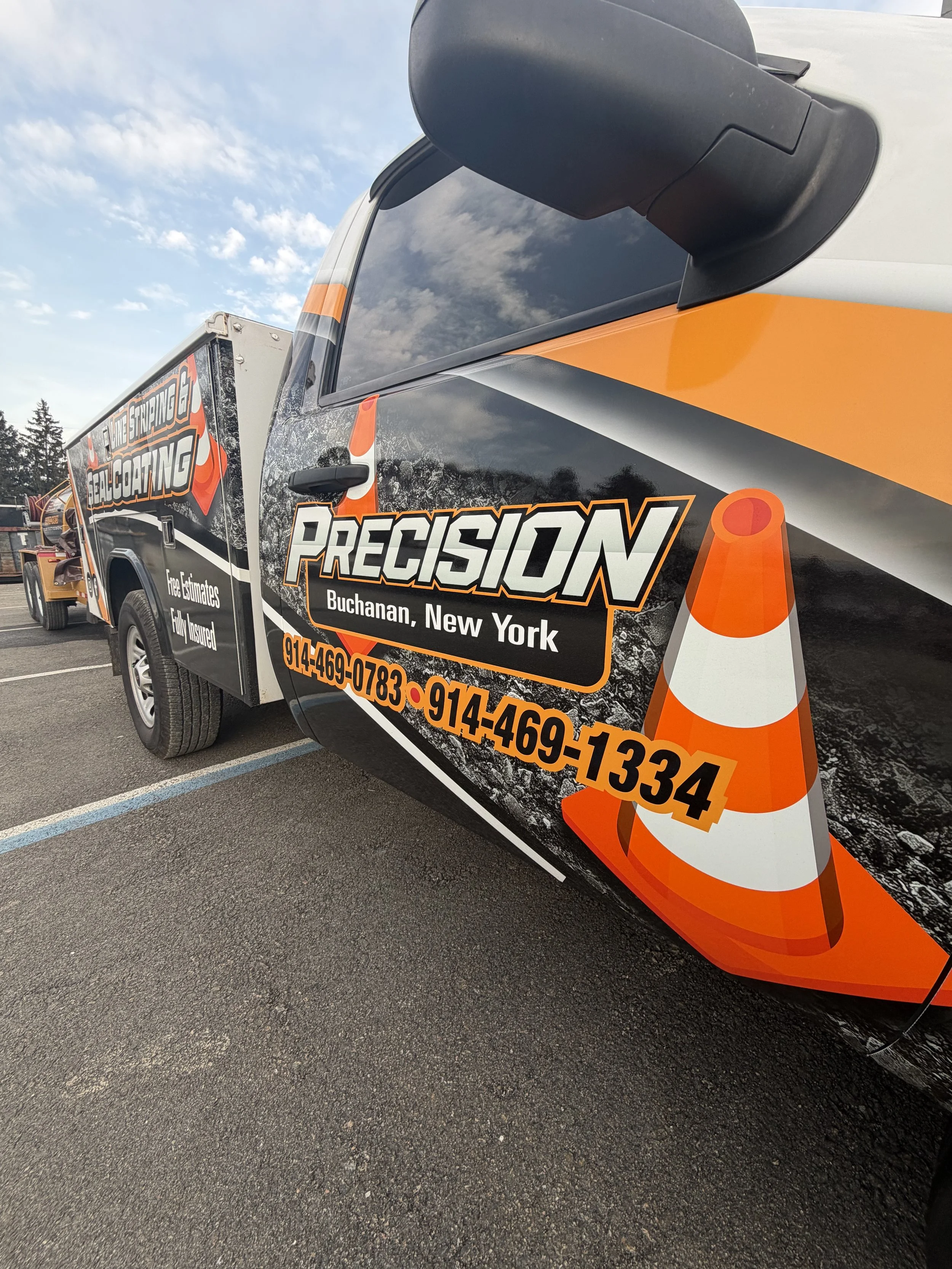 Side view of a black, orange, and white truck with the words 'Precision' and contact phone numbers, parked in a lot with other vehicles and a partly cloudy sky.