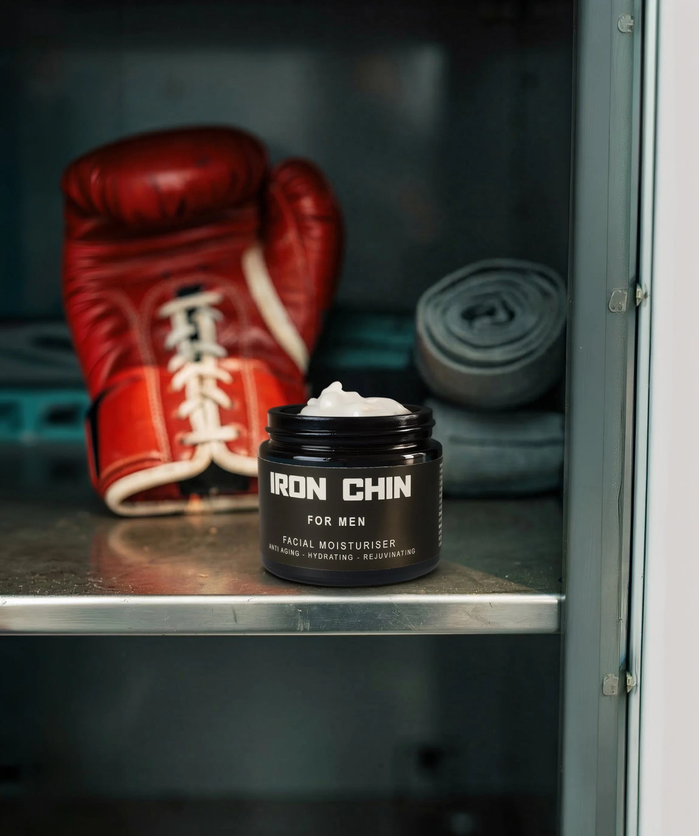 A black cosmetic jar labeled "Iron Chin for men facial moisturiser" in a gym / boxing gym locker, with a pair of red boxing gloves and hand wraps in the background.