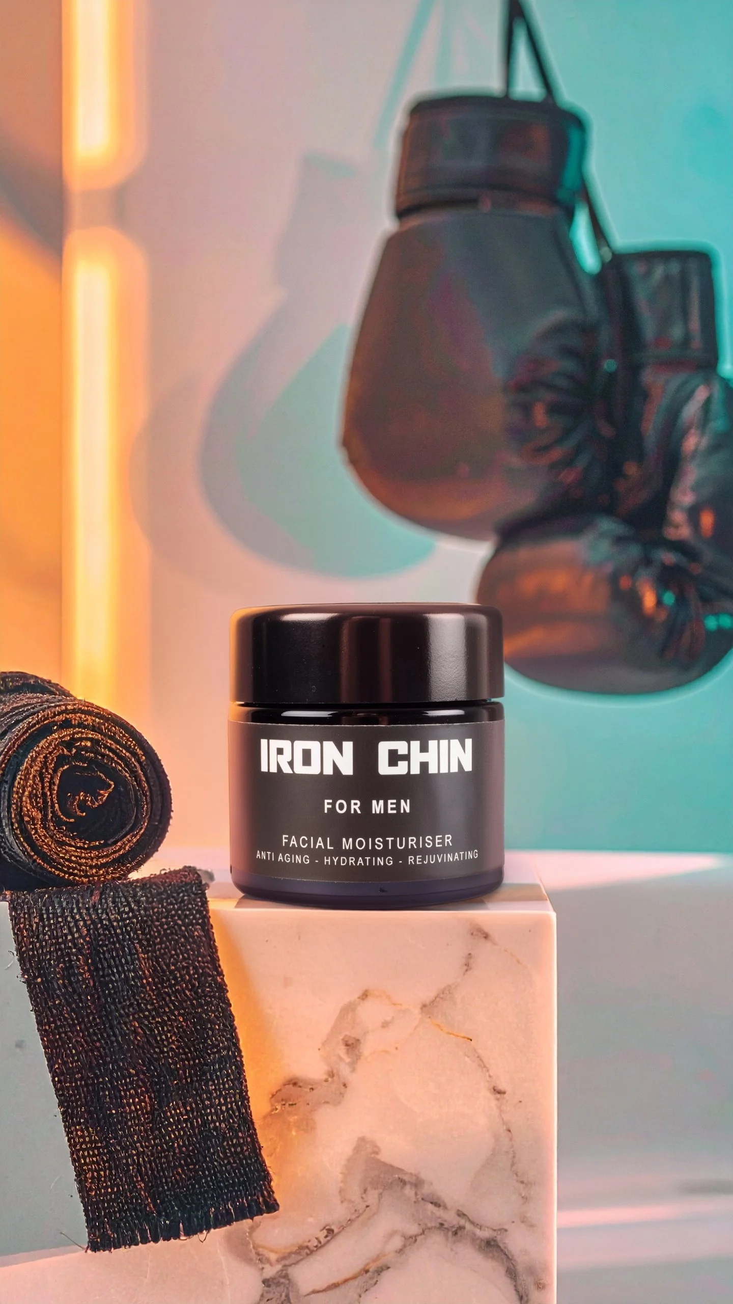 A black jar labeled 'Iron Chin for Men Facial Moisturizer' sitting on a marble block, with martial arts hand wraps to the left and a colorful abstract background featuring a pair of boxing gloves.