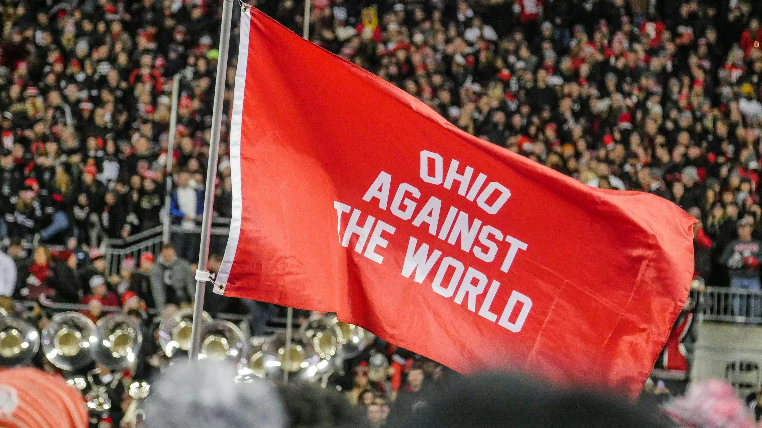 A red flag with white text that reads 'OHI O AGAINST THE WORLD' being held up at a crowded outdoor event. There are many people in the background and brass instruments on the ground.
