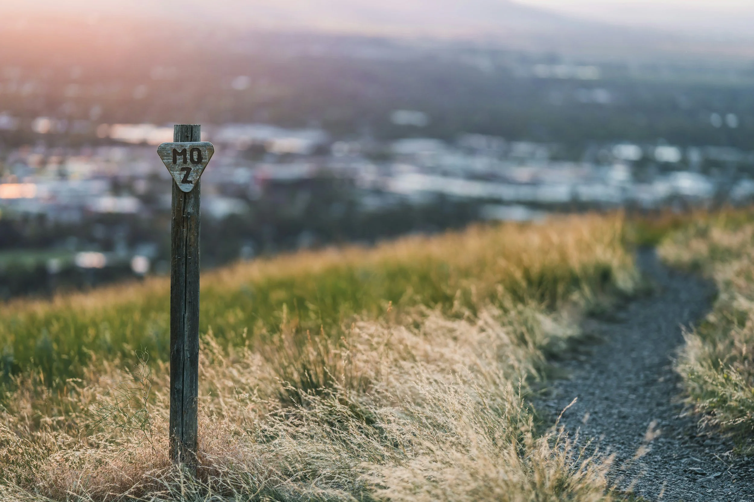 A wooden post with a metal plaque showing the number 'M 0' on a hillside overlooking a blurred cityscape in the distance during sunset.